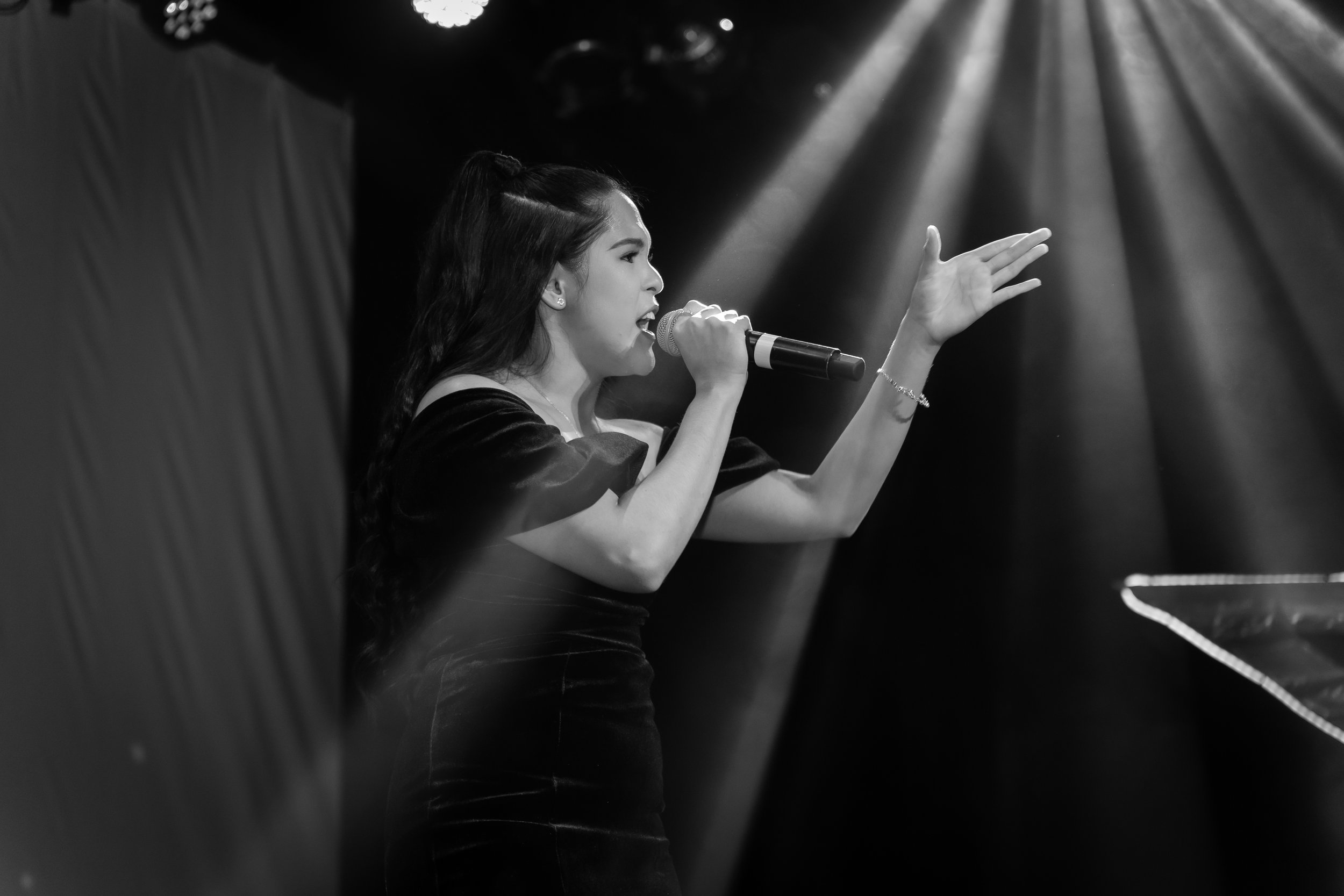 A woman singing on stage into a microphone, with her right hand raised and unless body language, under focused stage lighting.