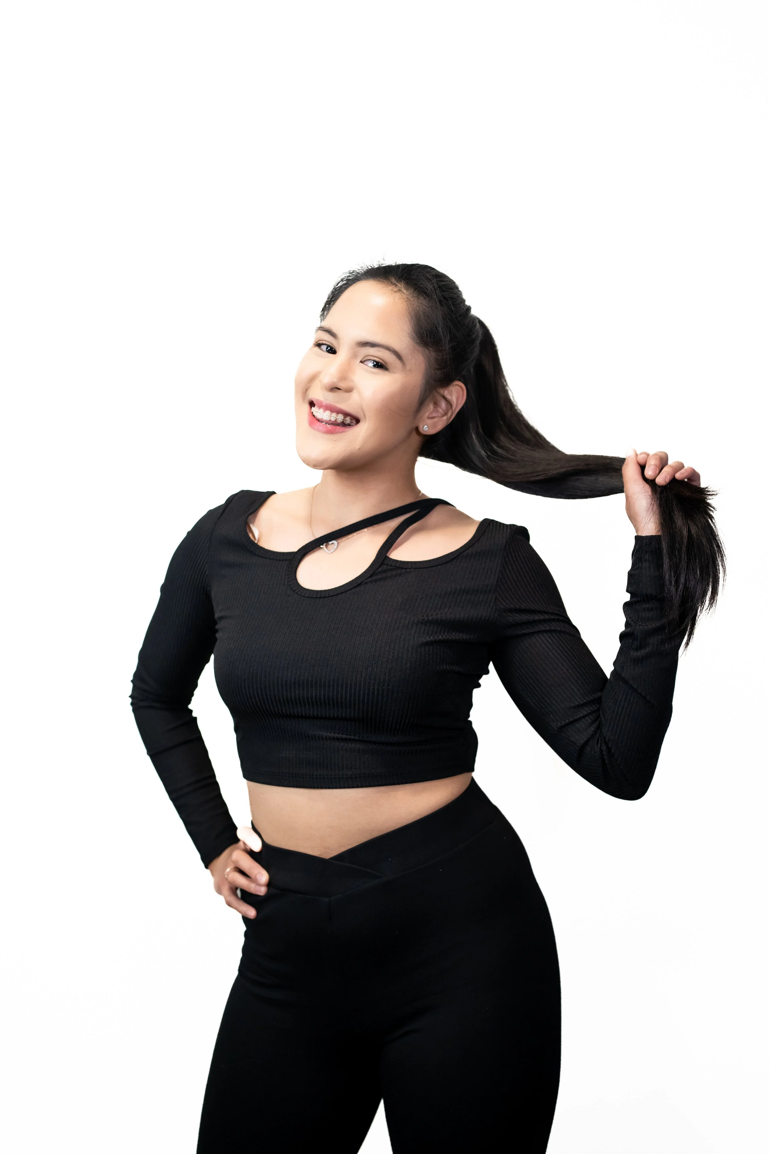 A young woman with long black hair, smiling and holding her hair with her right hand, wearing a black long-sleeve crop top and black pants against a plain white background.