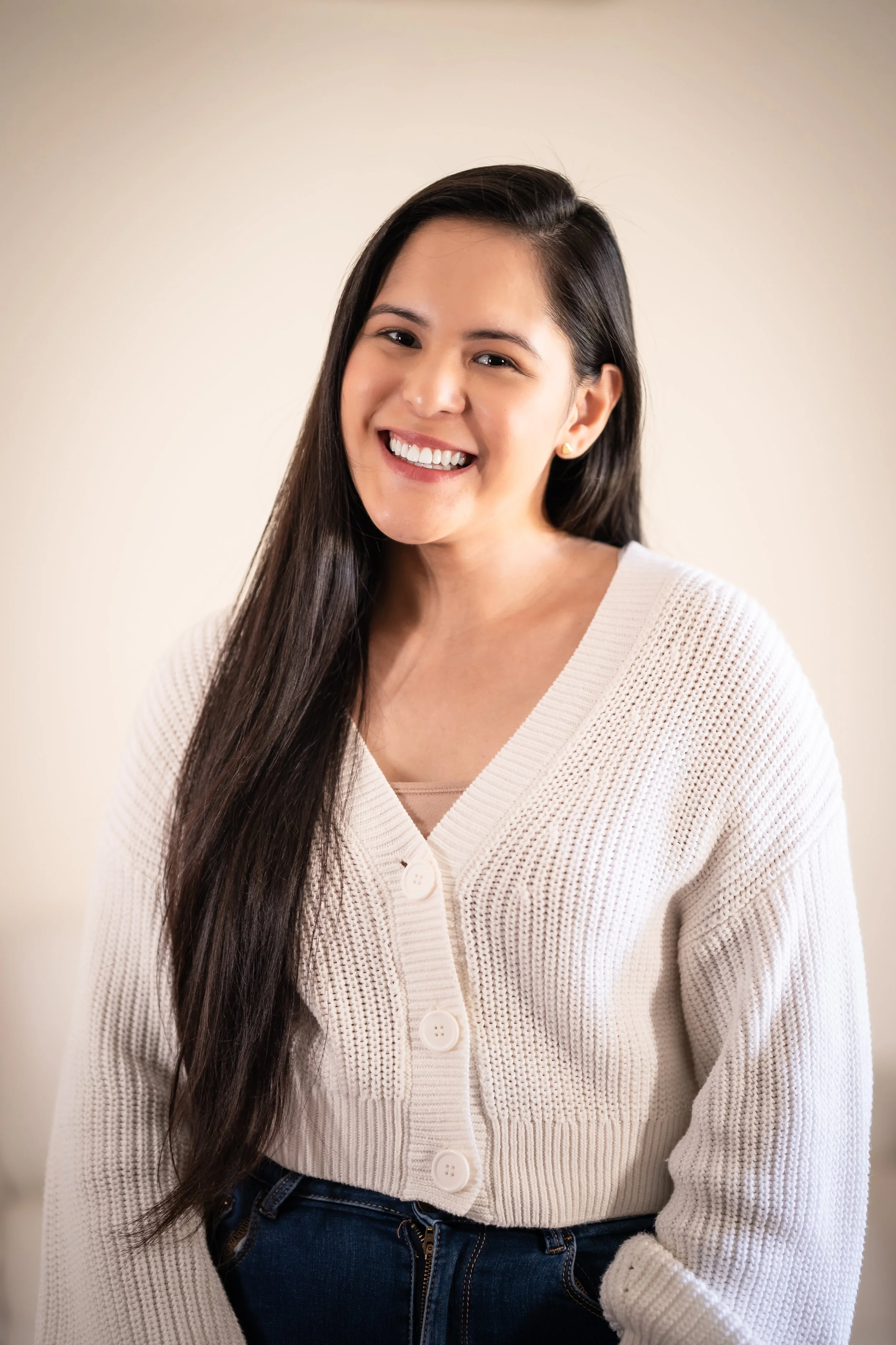 Young woman smiling with long dark hair, wearing a white knit cardigan and dark jeans, standing against a neutral background.