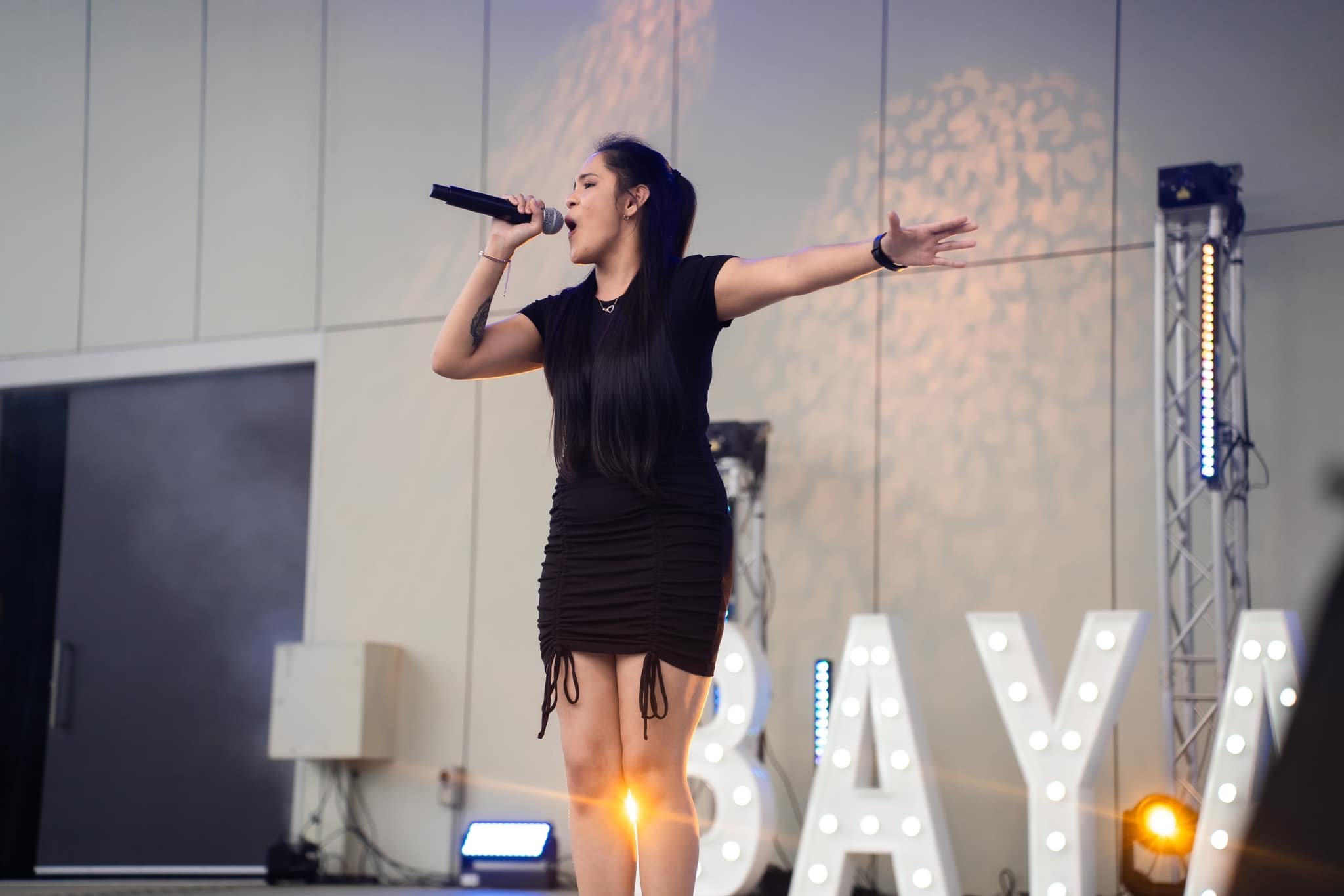 Young woman with long dark hair singing into a microphone on stage, with her left arm extended outward, in front of illuminated letters spelling 'SAY' at an indoor event.