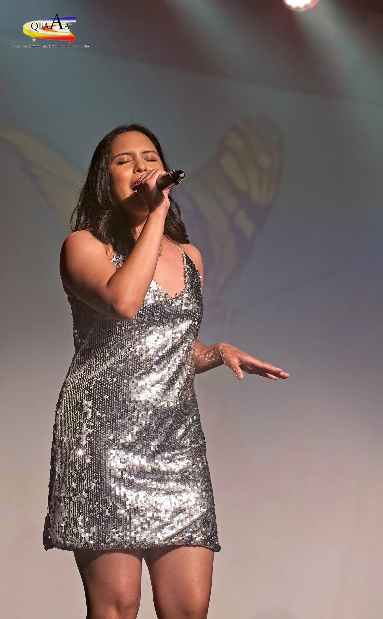 A woman in a silver sequin dress singing into a microphone on stage with a backdrop of the Queensland Filipino Australian Arts Association logo.