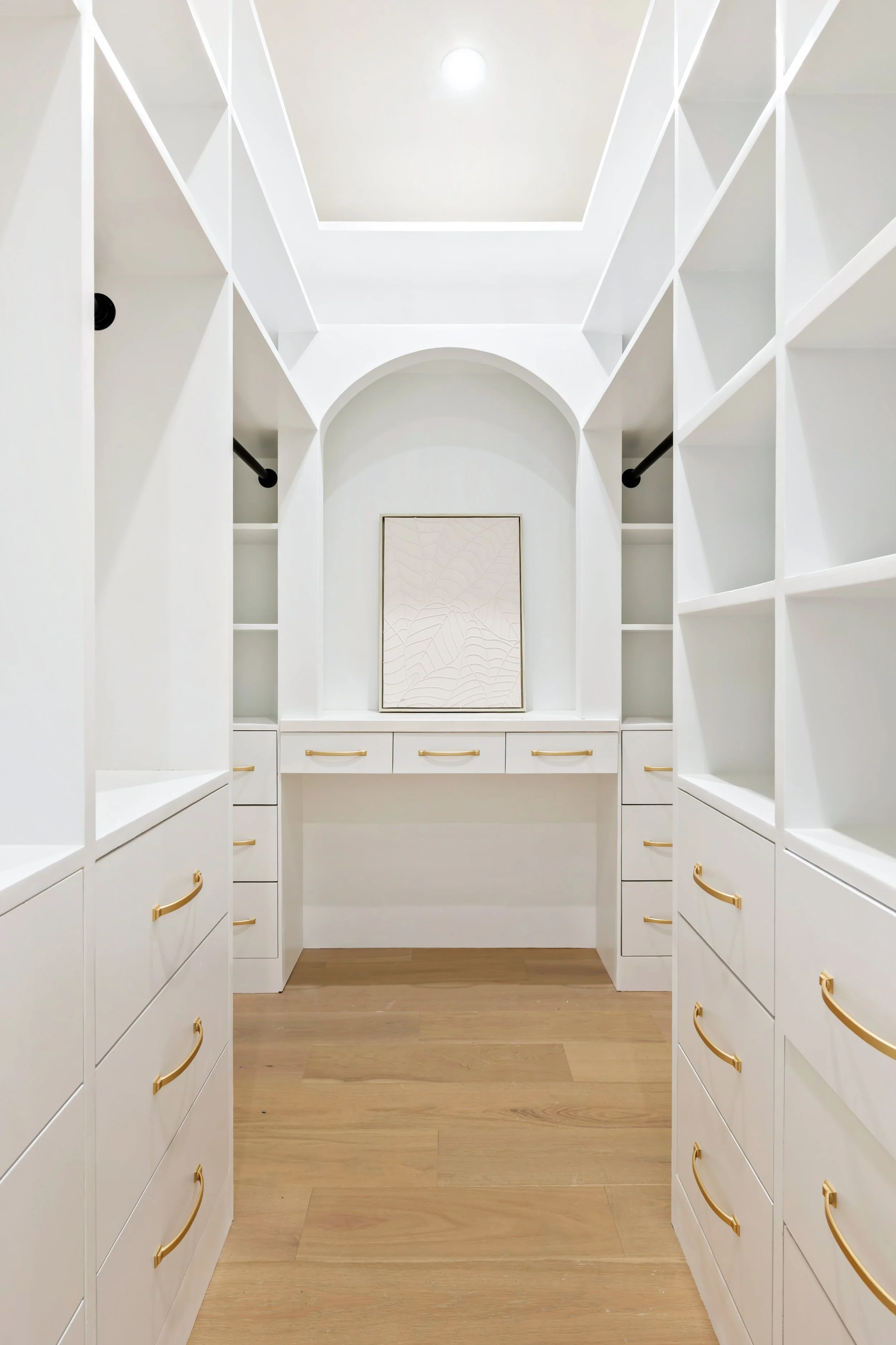 Primary closet with white custom cabinetry, brass hardware, and arched vanity space.