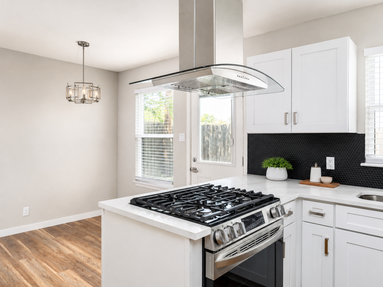 Kitchen with white cabinetry, modern statement glass vent hood, gas cooktop with stainless steel range, black backsplash detail and transitional brushed nickel and glass pendant light.