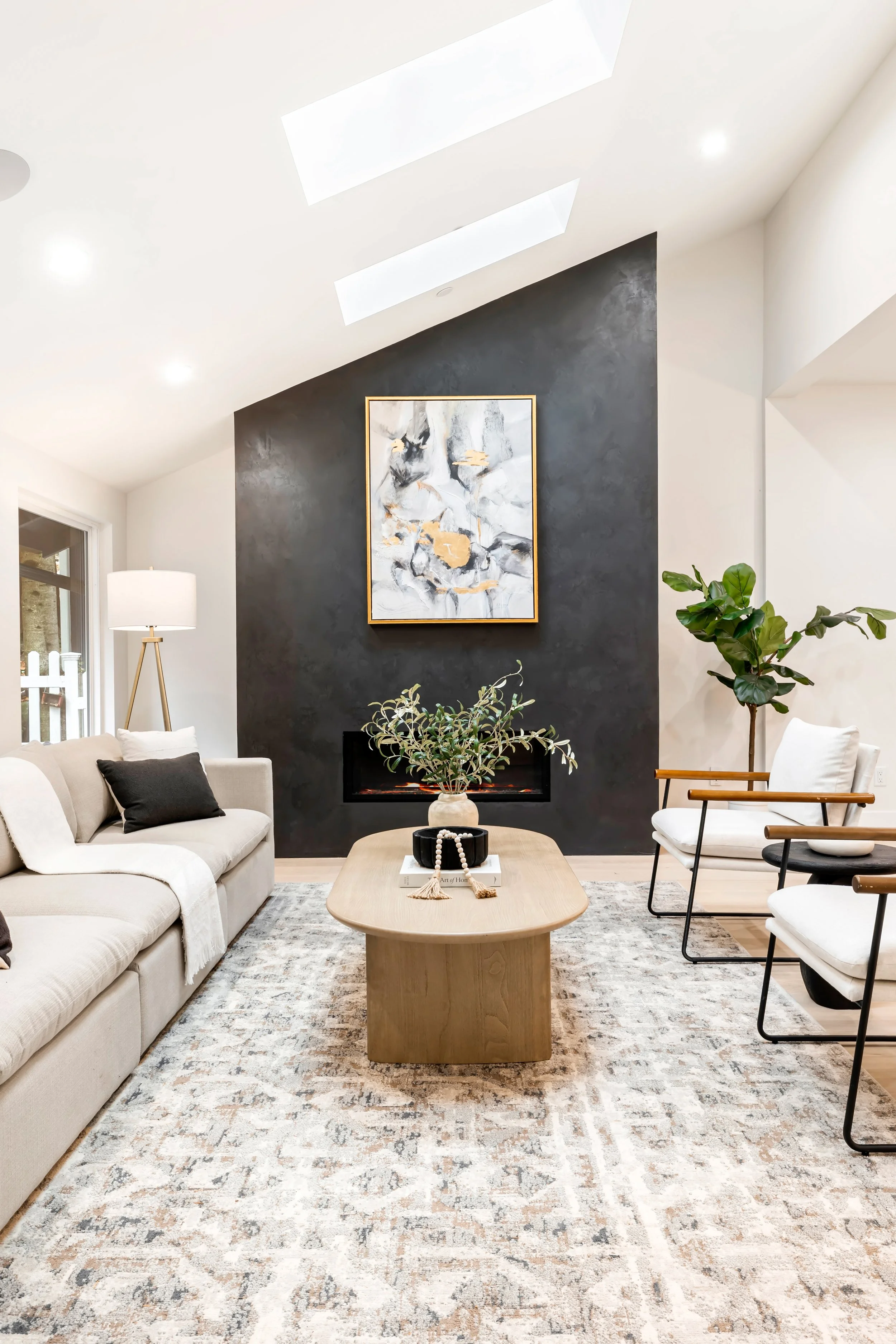 Contemporary living room with black asymmetrical plaster fireplace accent wall, white ceiling with skylights, neutral furniture, and wood coffee table.
