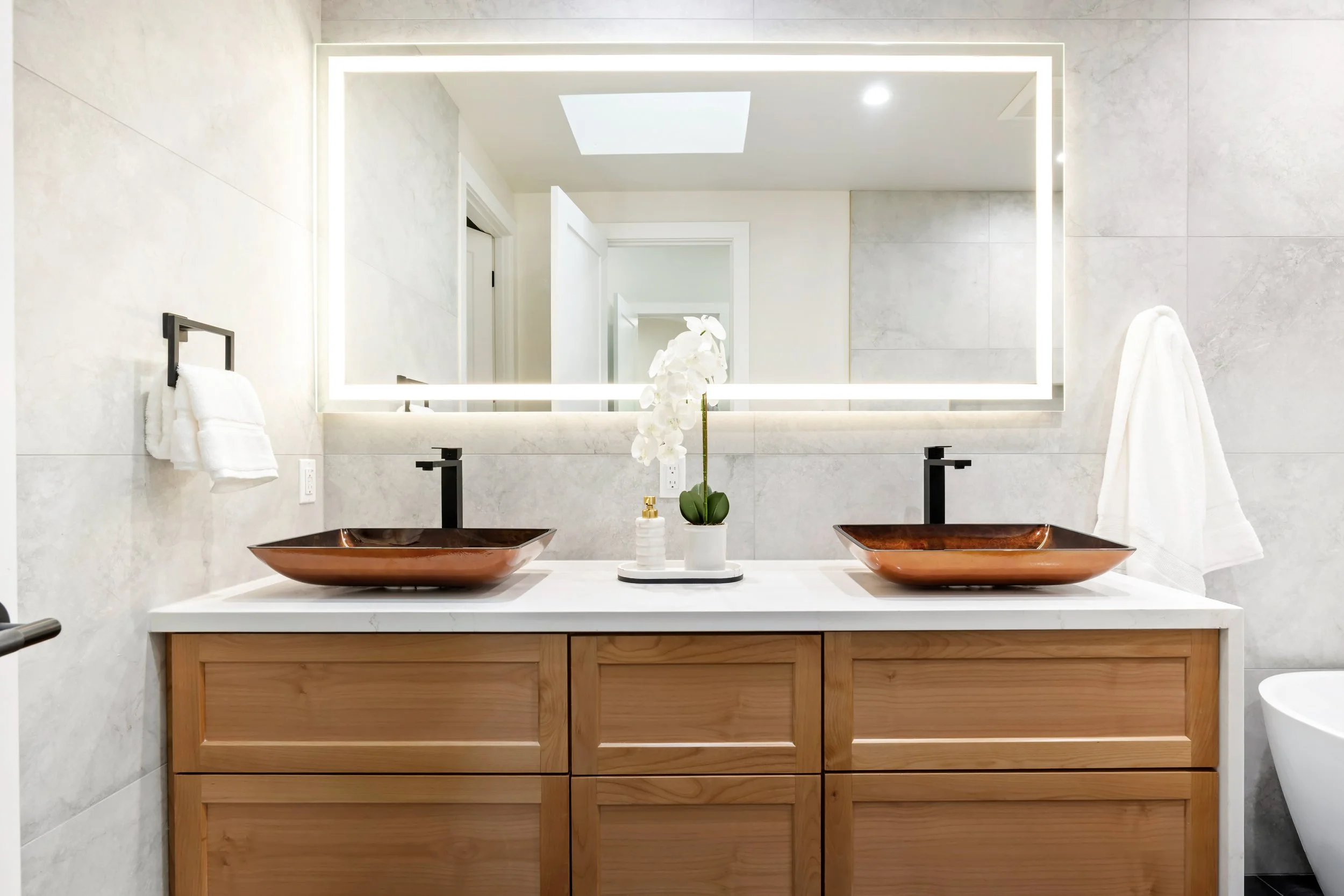 Primary bathroom vanity with marble tile, wood cabinetry, copper-toned glass vessel sinks and extra large illuminated mirror.
