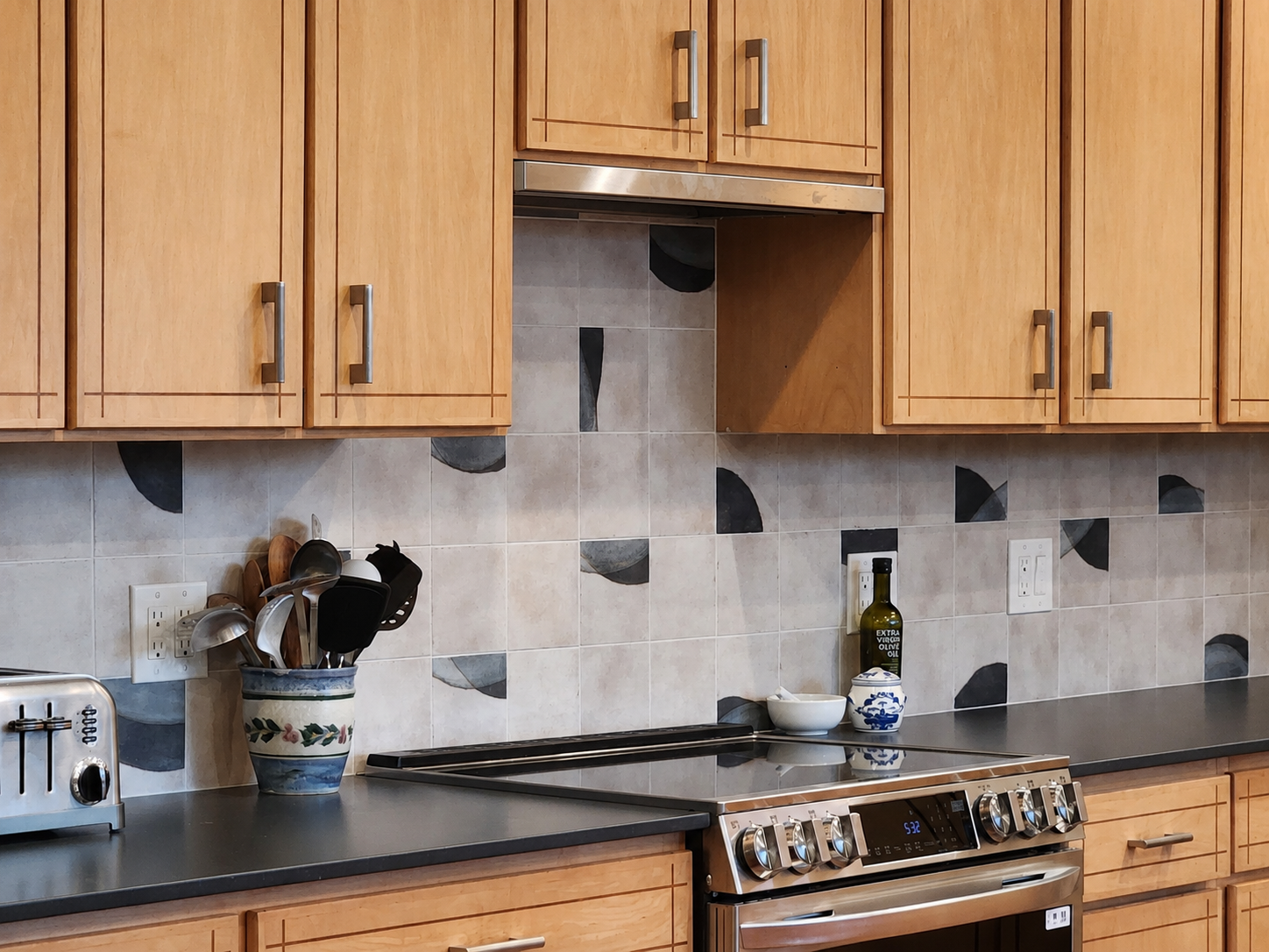 Kitchen cooking area with soapstone countertop, patterned tile backsplash, custom wood cabinetry, and modern range hood.