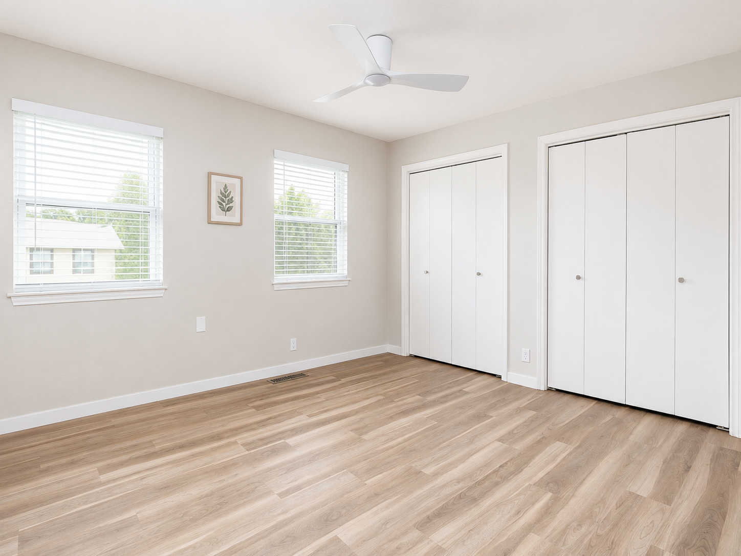 Bedroom with wood flooring, white closet doors, white ceiling fan, natural light from windows, and neutral wall color.