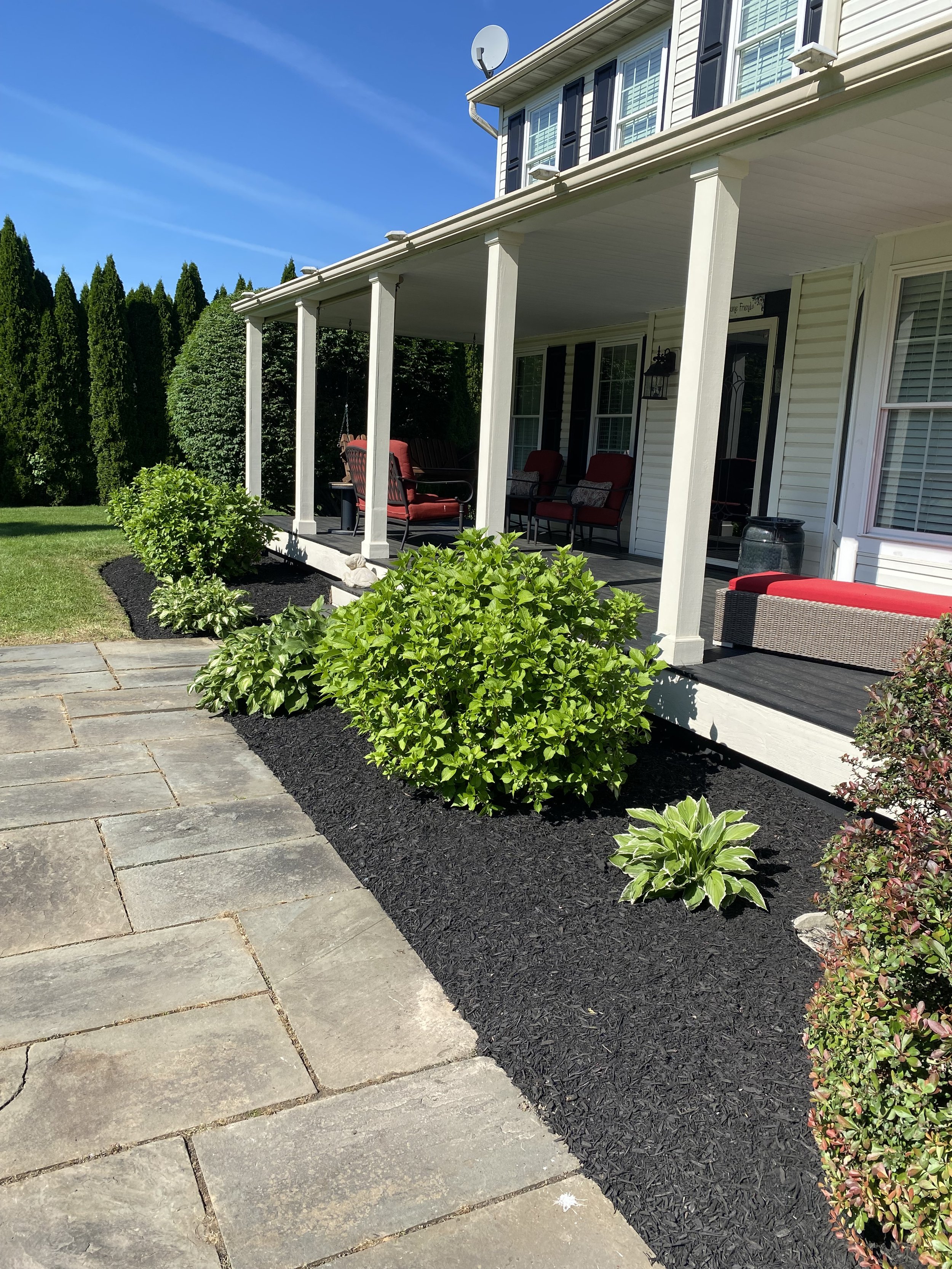 A suburban house with a covered porch, featuring outdoor seating with red cushions and a garden with green shrubs and mulch, alongside a stone pathway.