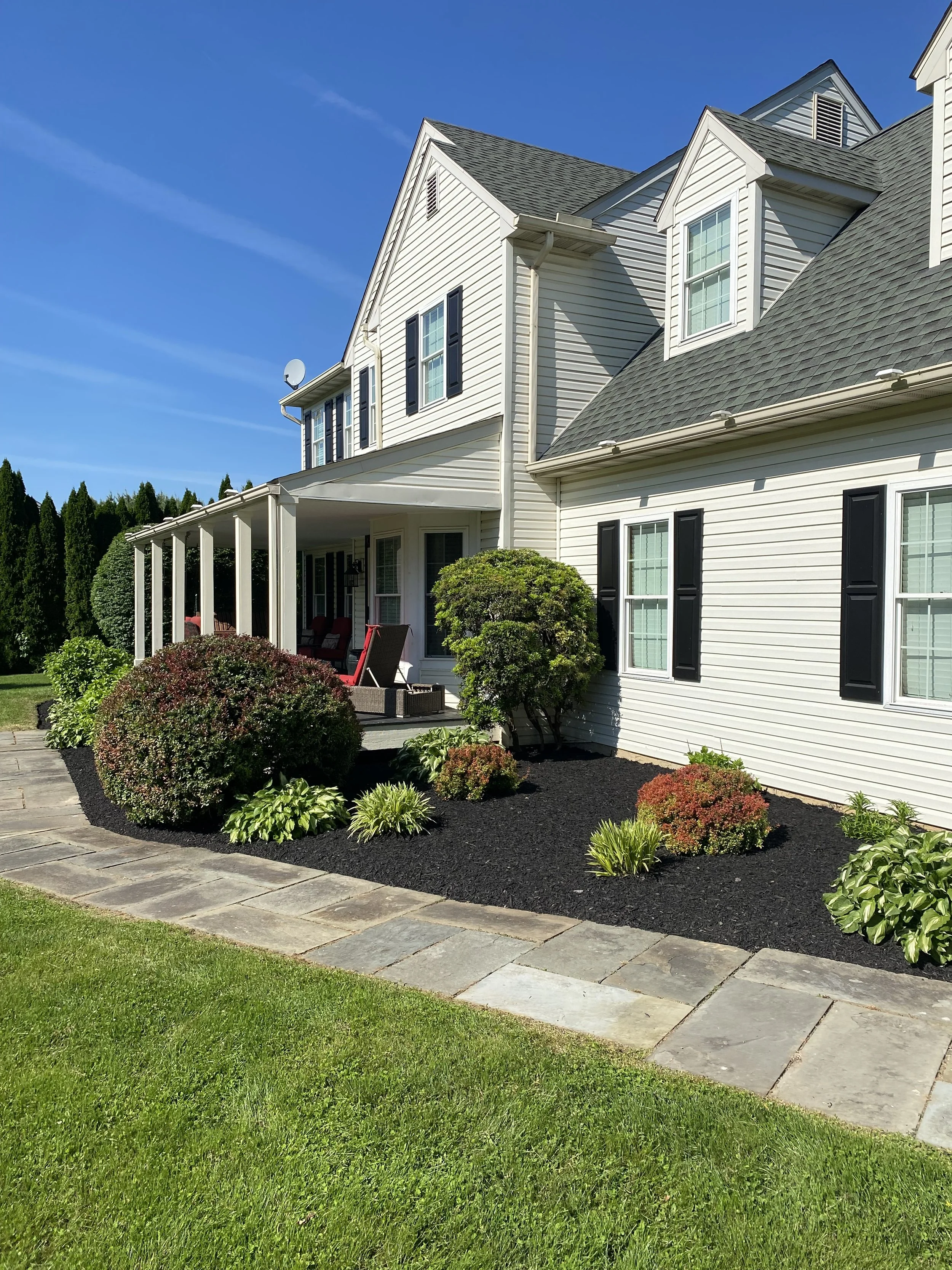 Front yard of a white two-story house with black shutters, a porch with red chairs, green shrubs, a stone pathway, and a manicured lawn under a clear blue sky.
