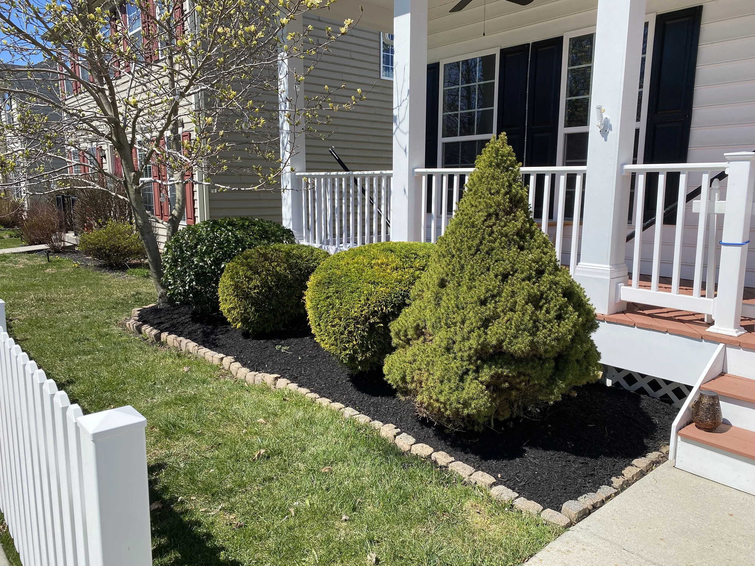 A front yard garden with various green bushes and a small tree, bordered by brick edging, in front of a house with a white porch and stairs, with a lantern on the step.