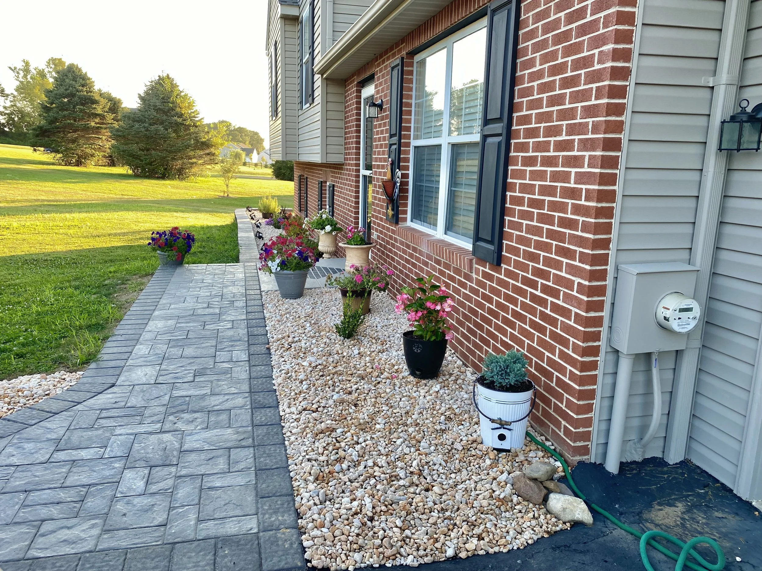 A paved walkway running along the side of a house with potted flowers and plants in flower pots of various sizes, placed on a bed of small white rocks. The house has brick and siding exterior with windows and blue shutters. There is a lush green lawn