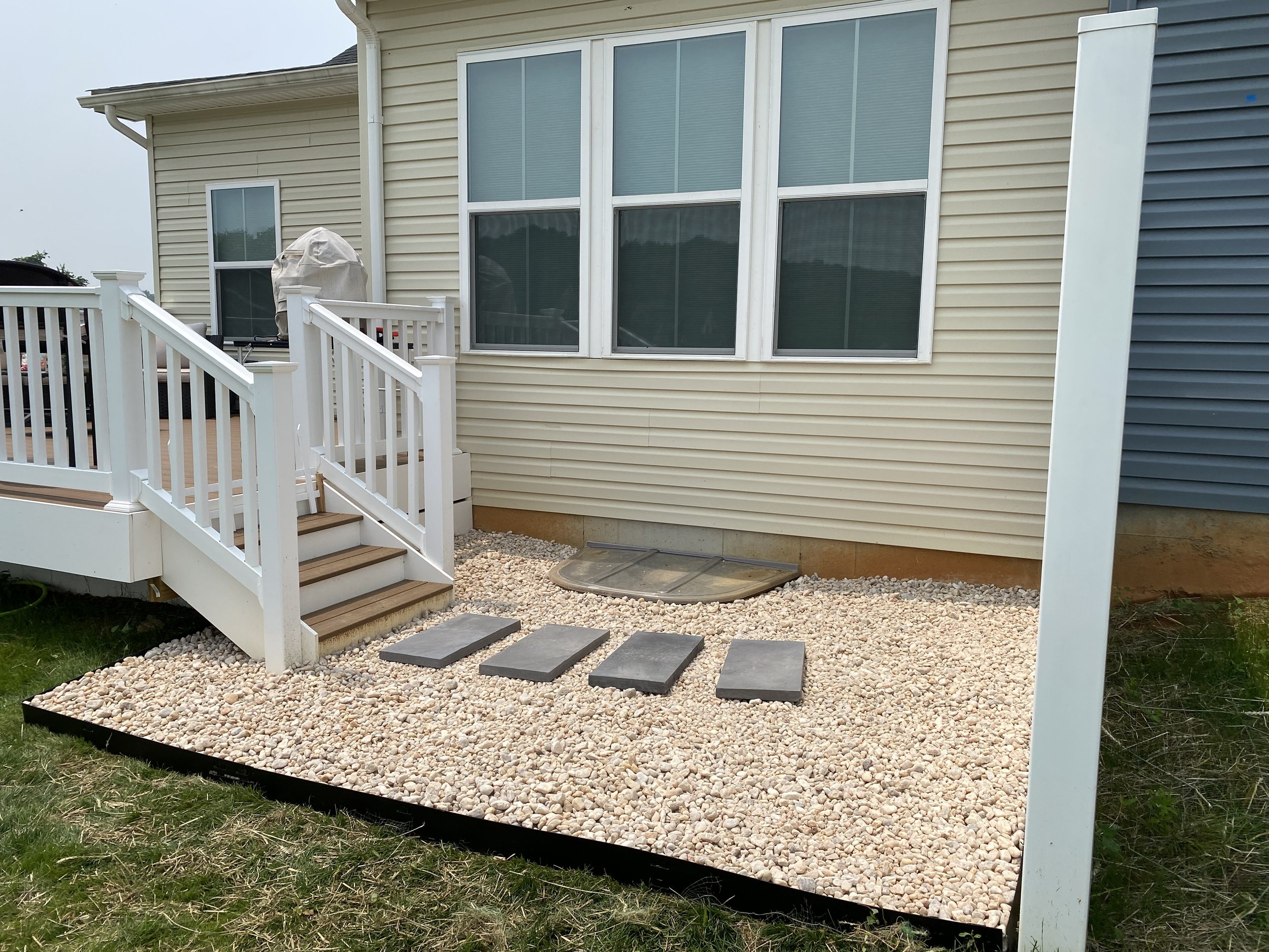 Backyard with a small deck, gravel ground, and three stepping stones leading to a screened door, with a house featuring beige siding and white-framed windows.