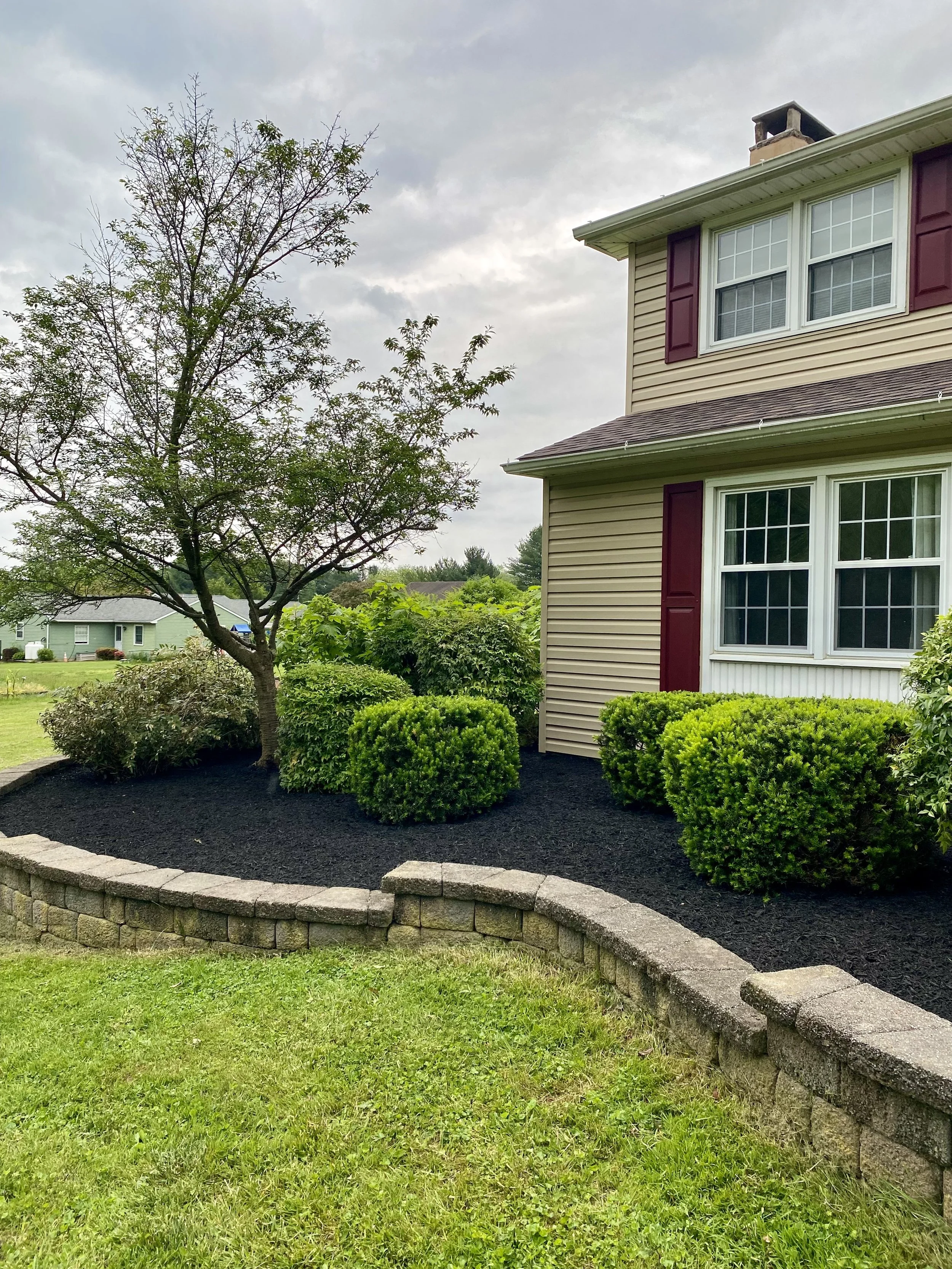 Front yard with a curved retaining wall made of stone blocks, green shrubs, a small tree, and a beige house with maroon shutters and white window frames. The sky is gray and cloudy.