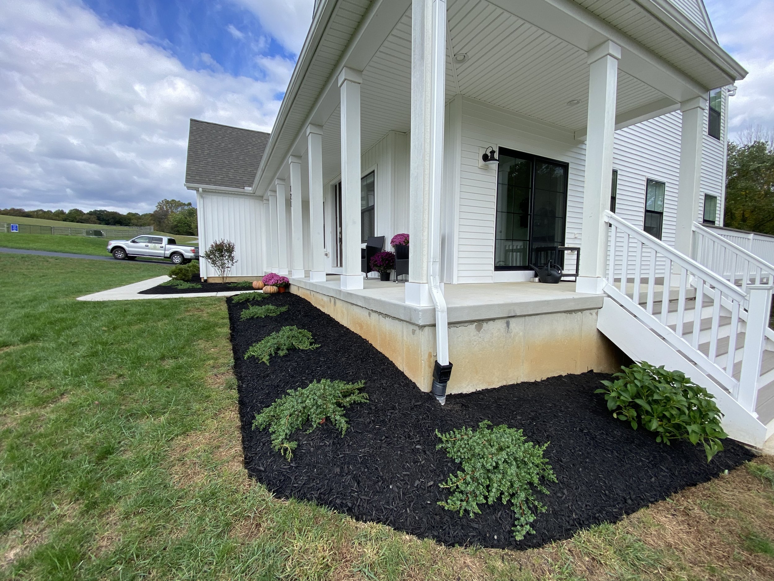 A white house with a porch, black mulch landscaping with small plants, and a sloped ramp with white railings leading to the front door. There is a lawn, a car in the driveway, and a partly cloudy sky.