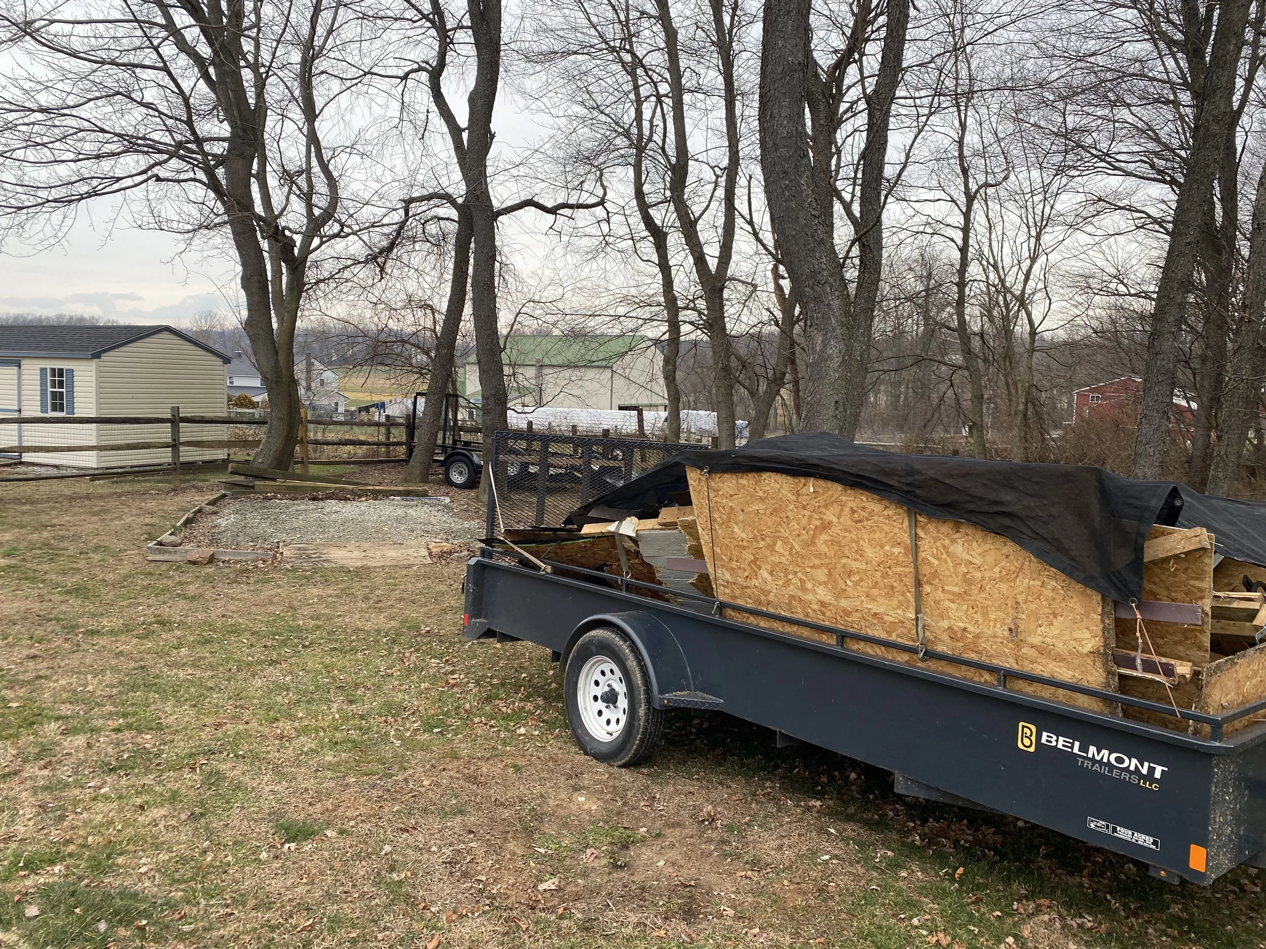 A yard with a trailer filled with lumber covered partly with a black tarp, a small area of gravel, and trees without leaves, indicating late fall or winter.