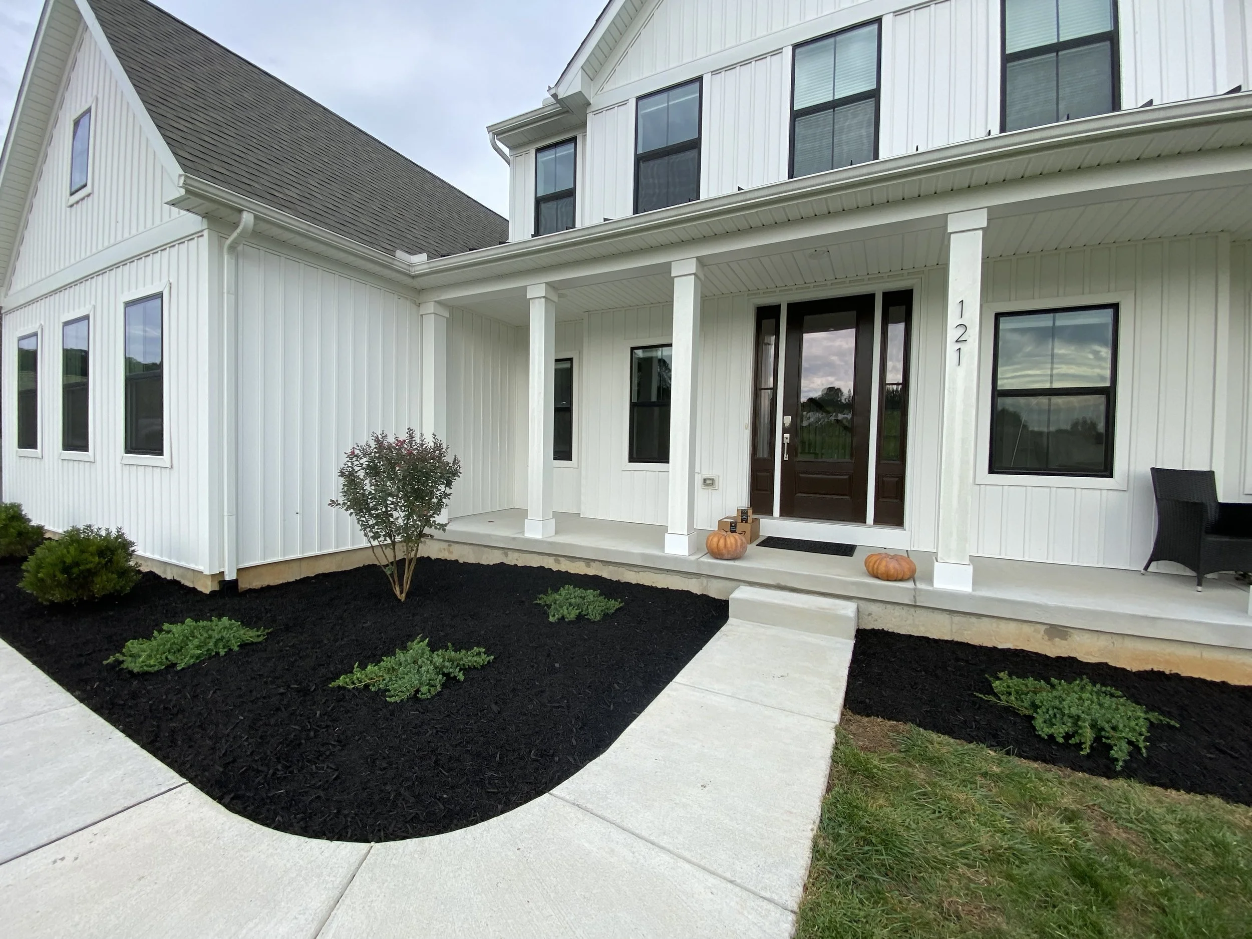 Front porch of a modern white house with black front door, pumpkins on steps, and numbered 121, landscaped with mulch and small plants.