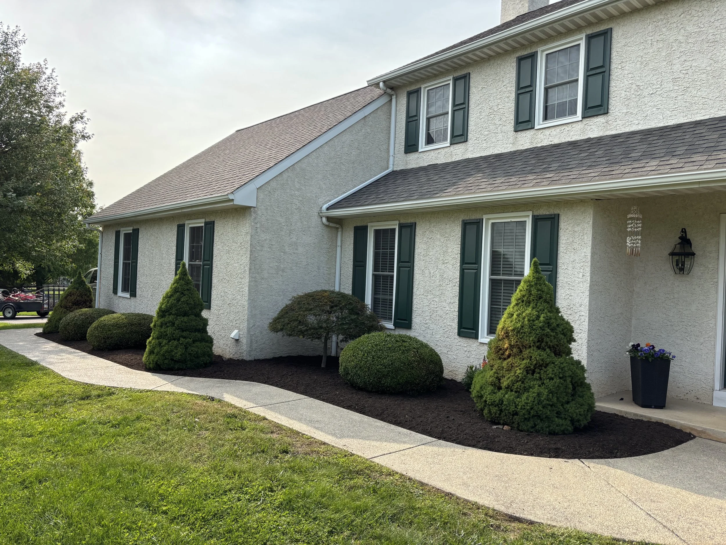 Front view of a house with white textured walls, dark green shutters, and a brown shingle roof. The house has multiple windows and a small porch with a flowerpot and outdoor lantern. The yard features neatly trimmed bushes and a curved concrete walkw