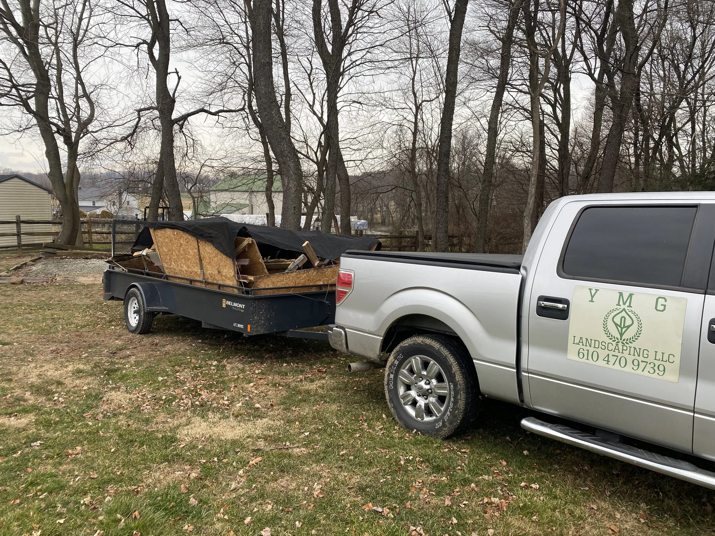 A silver pickup truck with a sign for Y M G Landscaping LLC parked on grass, hauling a black trailer loaded with debris, wood, and a black tarp in a backyard with trees and a wooden fence.