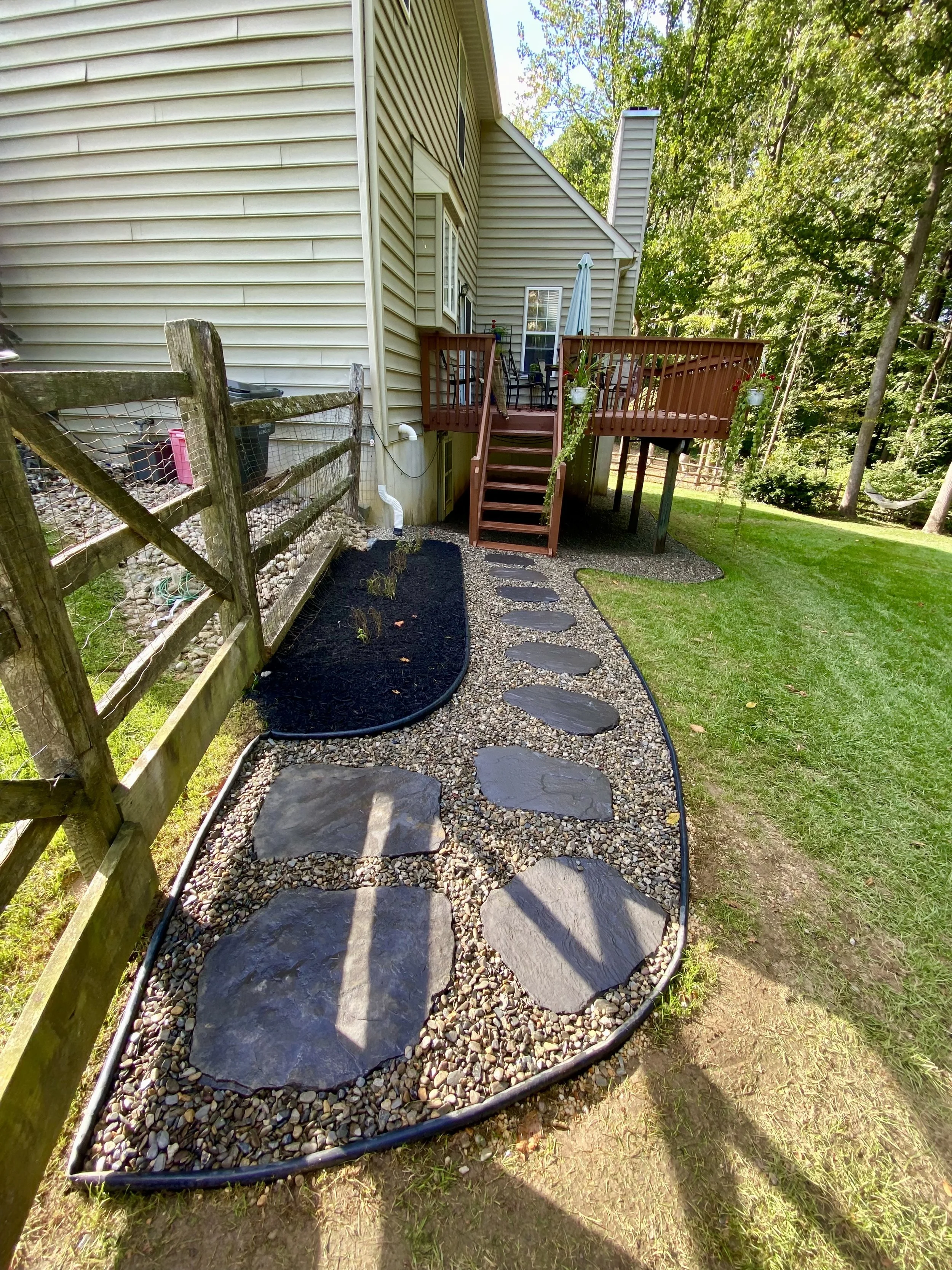 Backyard with stone pathway leading to a wooden deck, surrounded by green grass and trees.