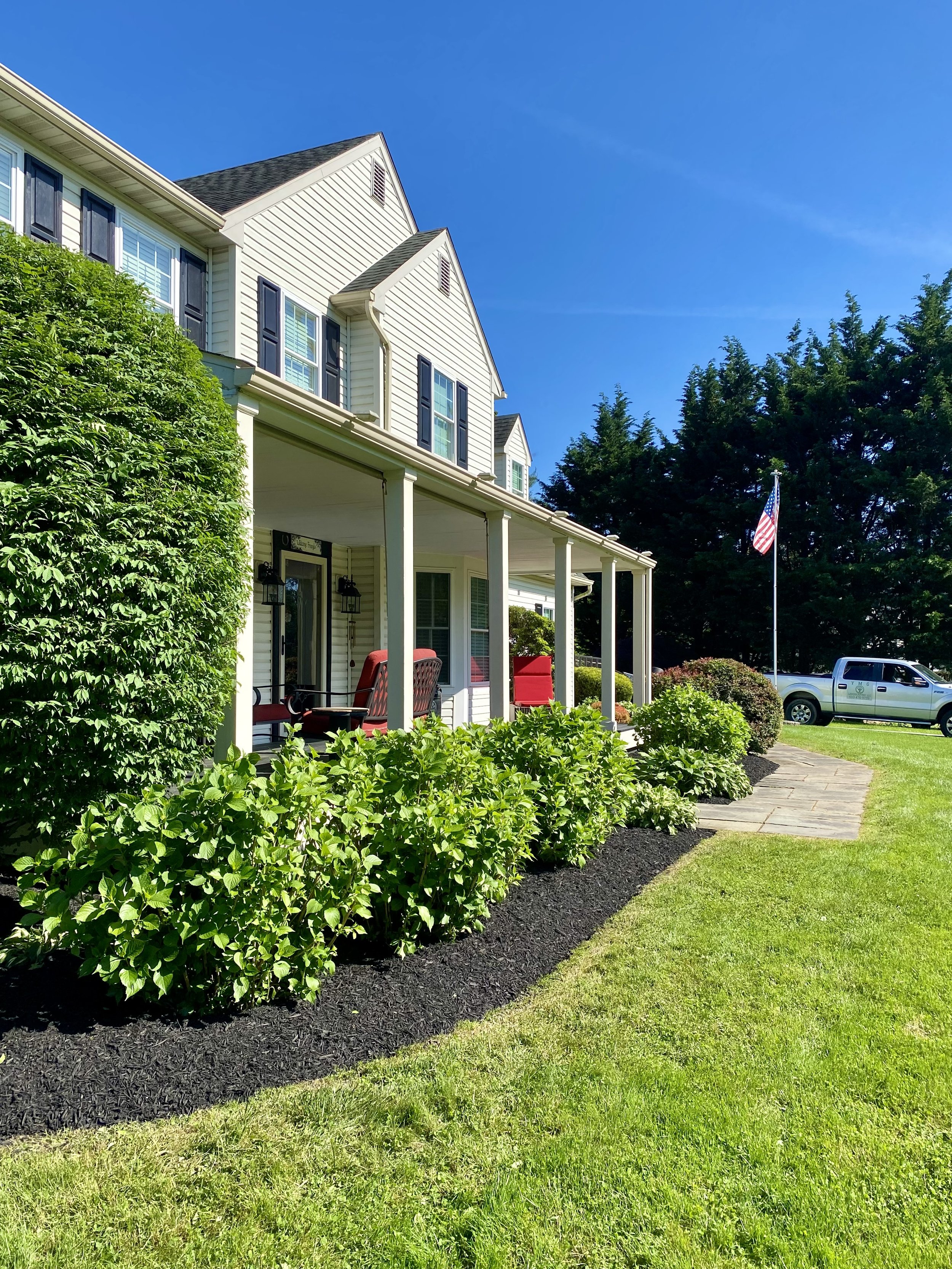 Front porch of a white two-story house with black shutters, Charleston-style columns, red outdoor furniture, a landscaped garden with green bushes, a stone pathway, an American flag, and a silver pickup truck in the background on a sunny day.