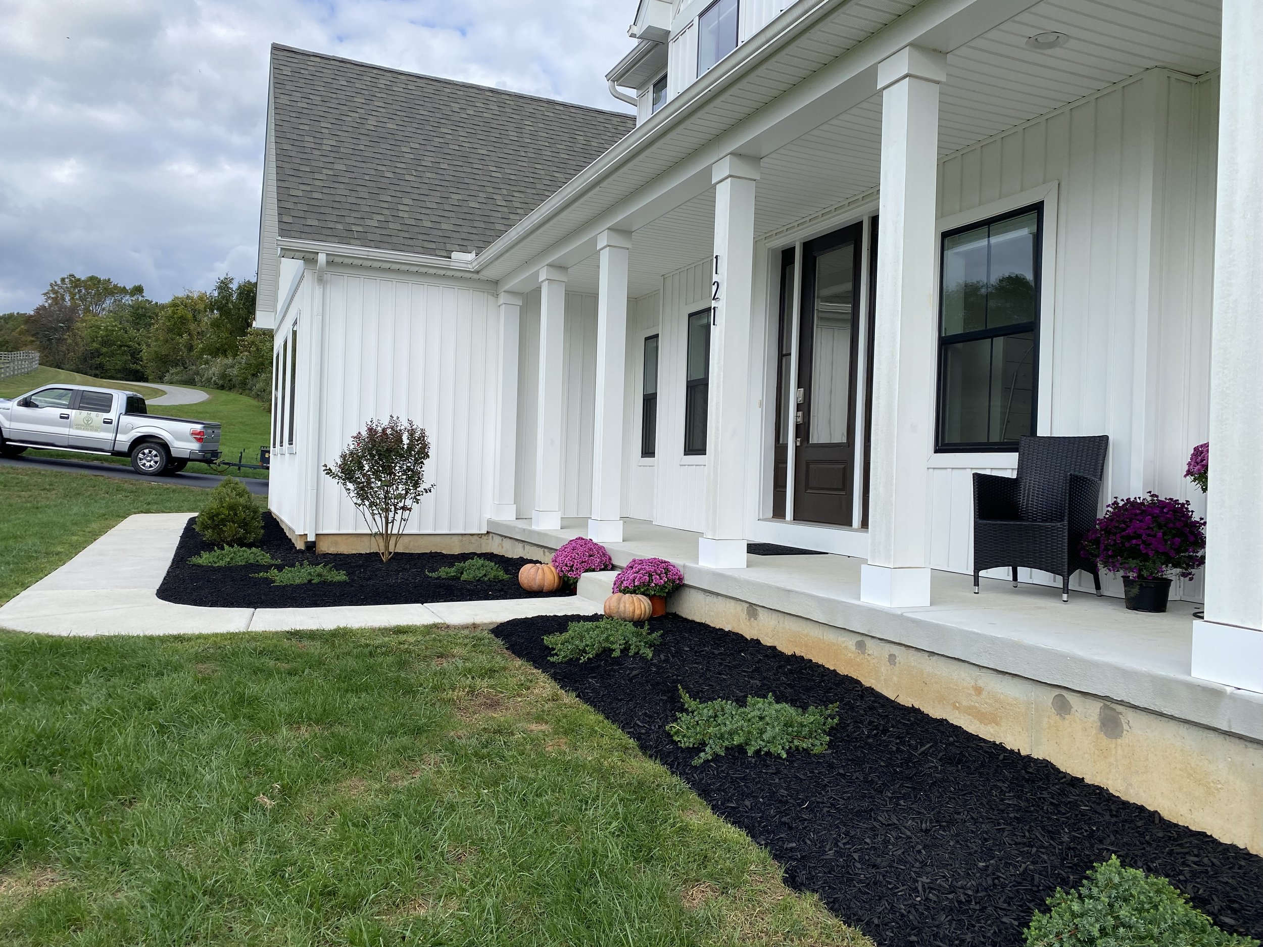 Front porch of a white house with black door, black chair, potted purple flowers, and yellow pumpkins, with landscaped black mulch garden beds, a grassy lawn, and a truck parked in the background.