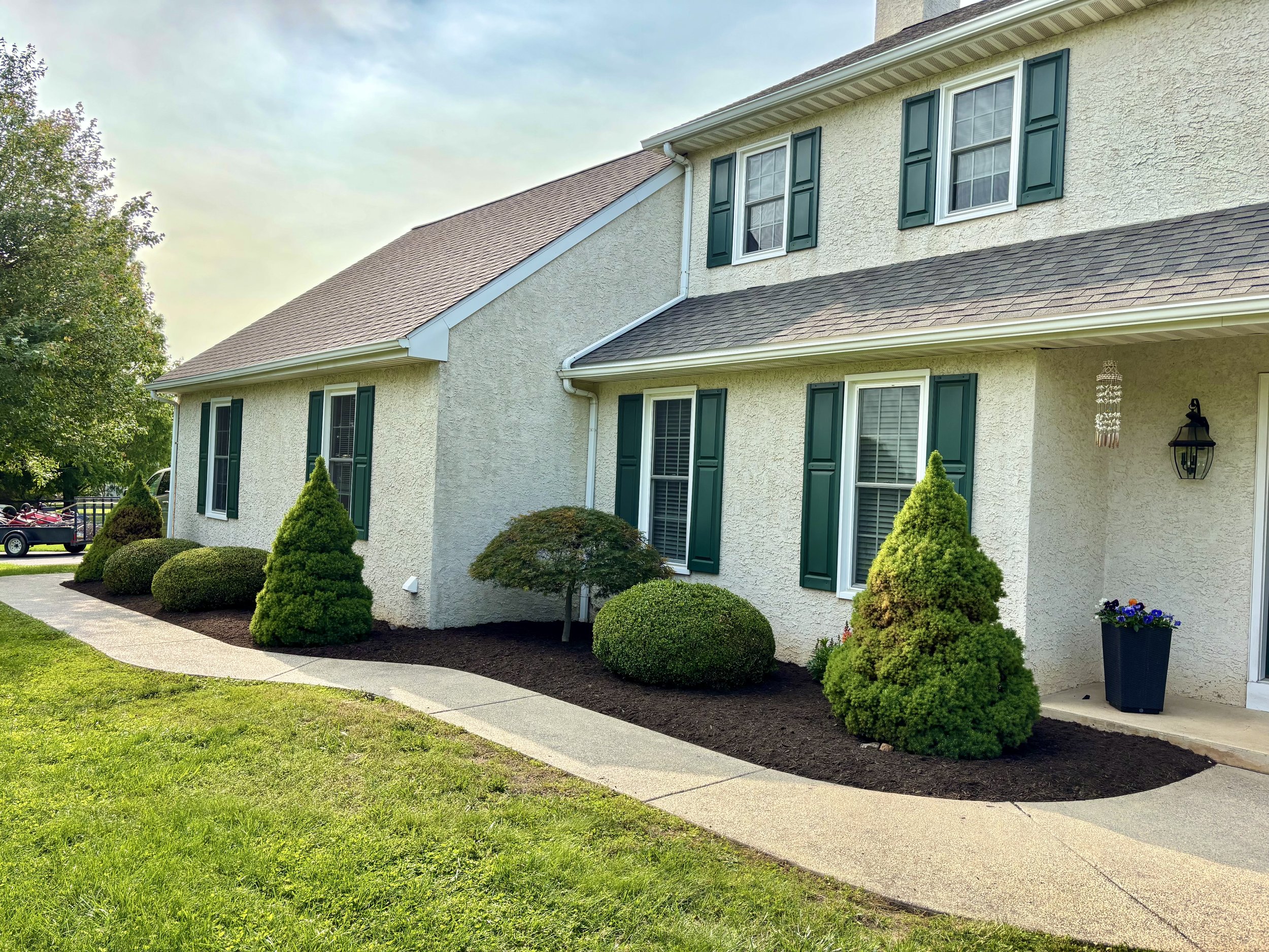 Front of a two-story house with white textured exterior walls, green shutters on windows, and a brown shingled roof. Well-maintained shrubbery lines the walkway leading to the entrance, with neatly trimmed bushes and a small tree. A black planter with flowers is near the front door, and a lantern-style light fixture is on the wall. A lawn extends in front of the house, and a vehicle with a trailer is visible in the background.
