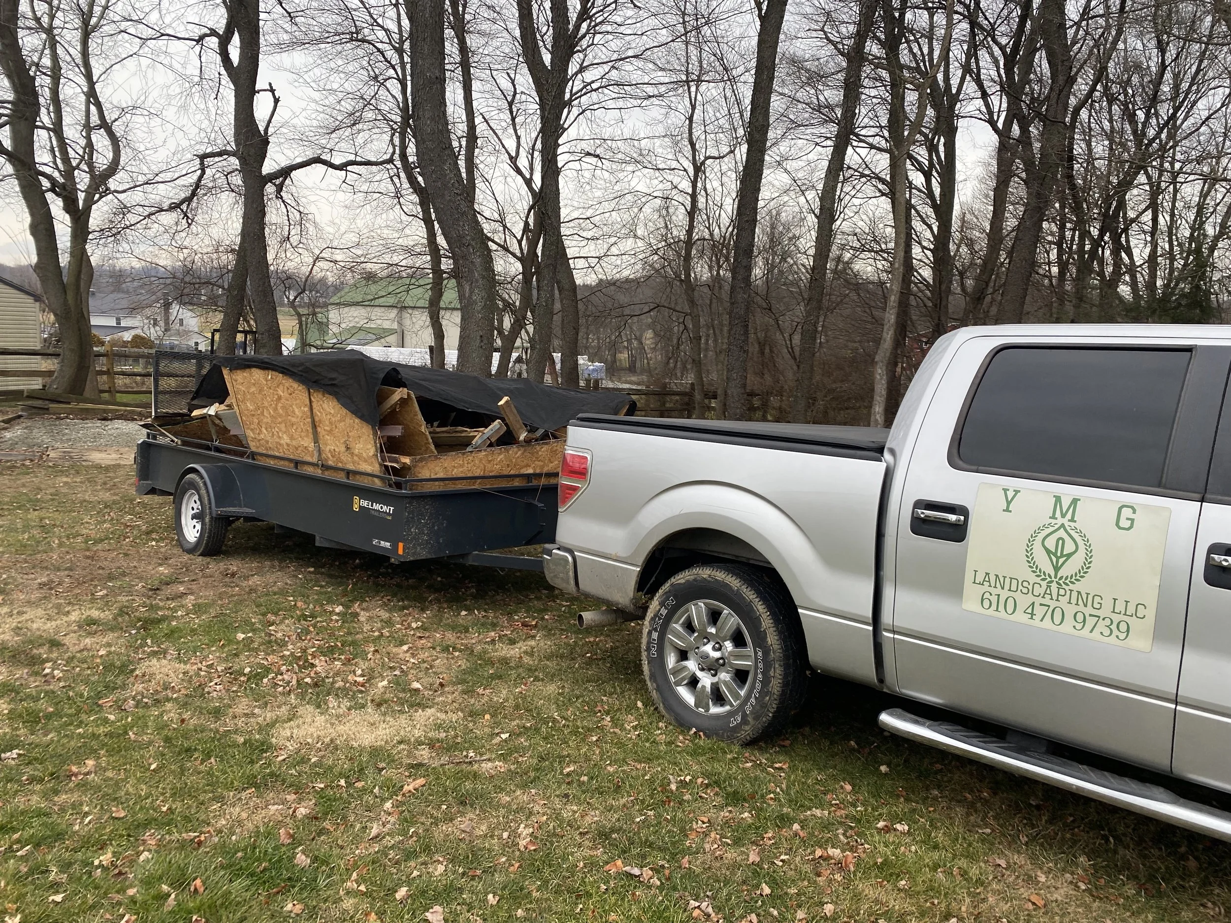 A pickup truck with a landscaping company logo parked on grass, towing a trailer filled with wood debris covered partially with a black tarp.