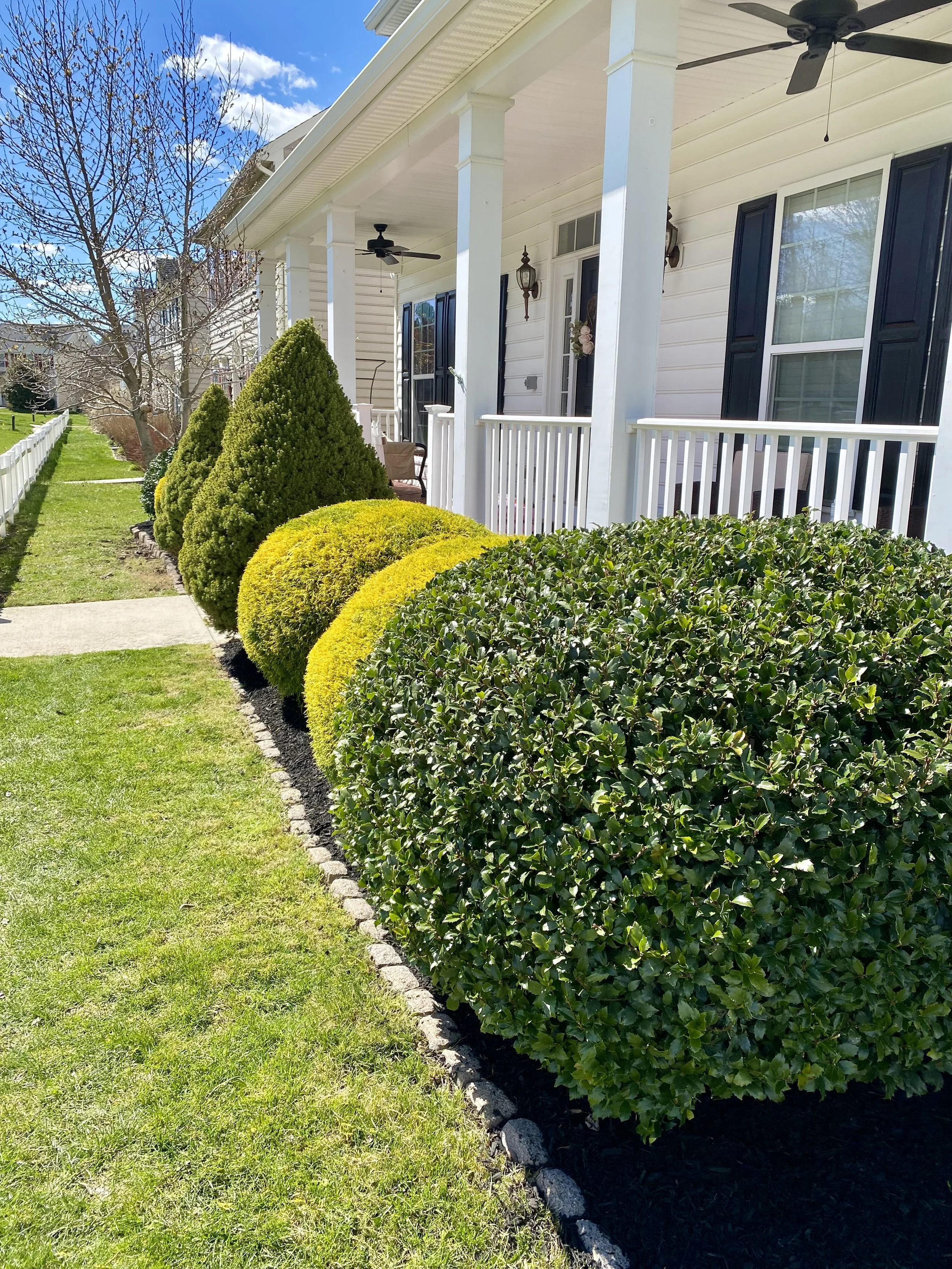 Front porch of a house with a white railing and dark shutters, lined with neatly trimmed bushes, and a sidewalk leading through a well-manicured lawn under a partly cloudy sky.