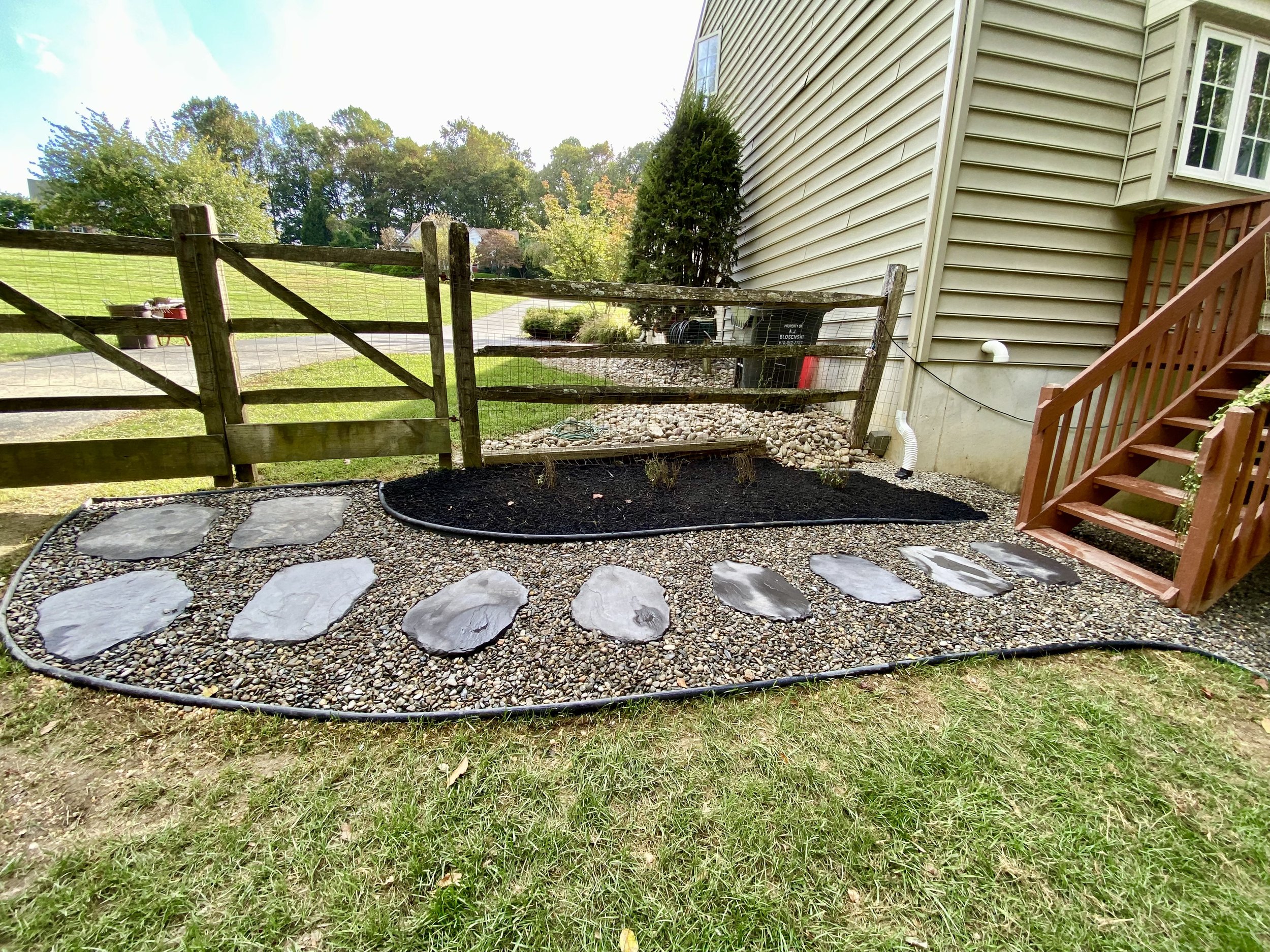 Backyard garden area with a stone pathway, freshly mulched flower bed, wooden staircase, and a wooden gate with a green yard and trees in the background.
