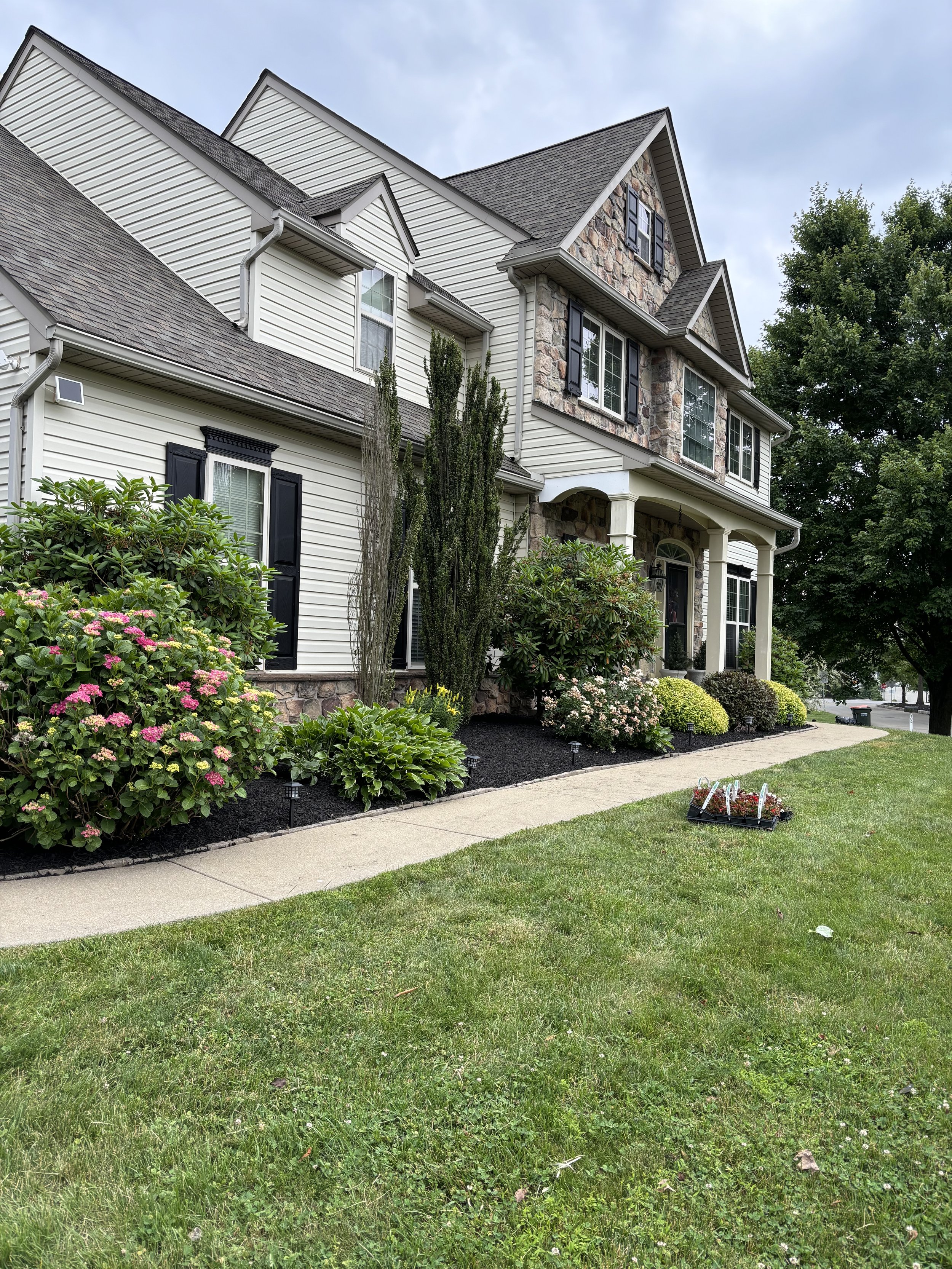Front yard of a large, multi-story house with beige siding and a stone facade. There are well-maintained flower beds with pink and white flowers, tall bushes, and trimmed shrubs. The house has black shutters, white columns, and a curved walkway leadi