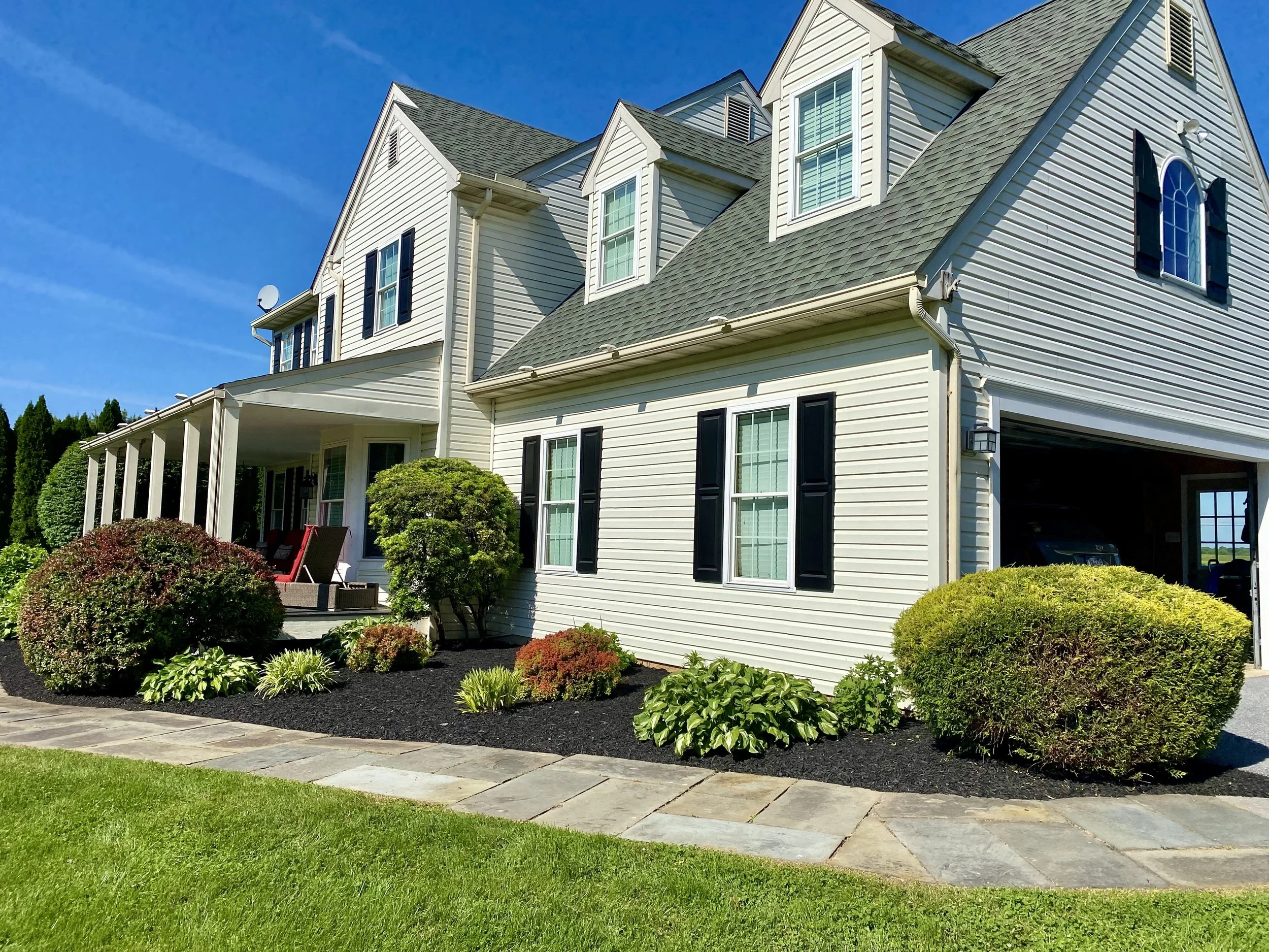 Exterior of a large suburban house with beige siding, black shutters, a green roof, and a front porch with outdoor seating, surrounded by well-maintained shrubbery and a stone walkway.