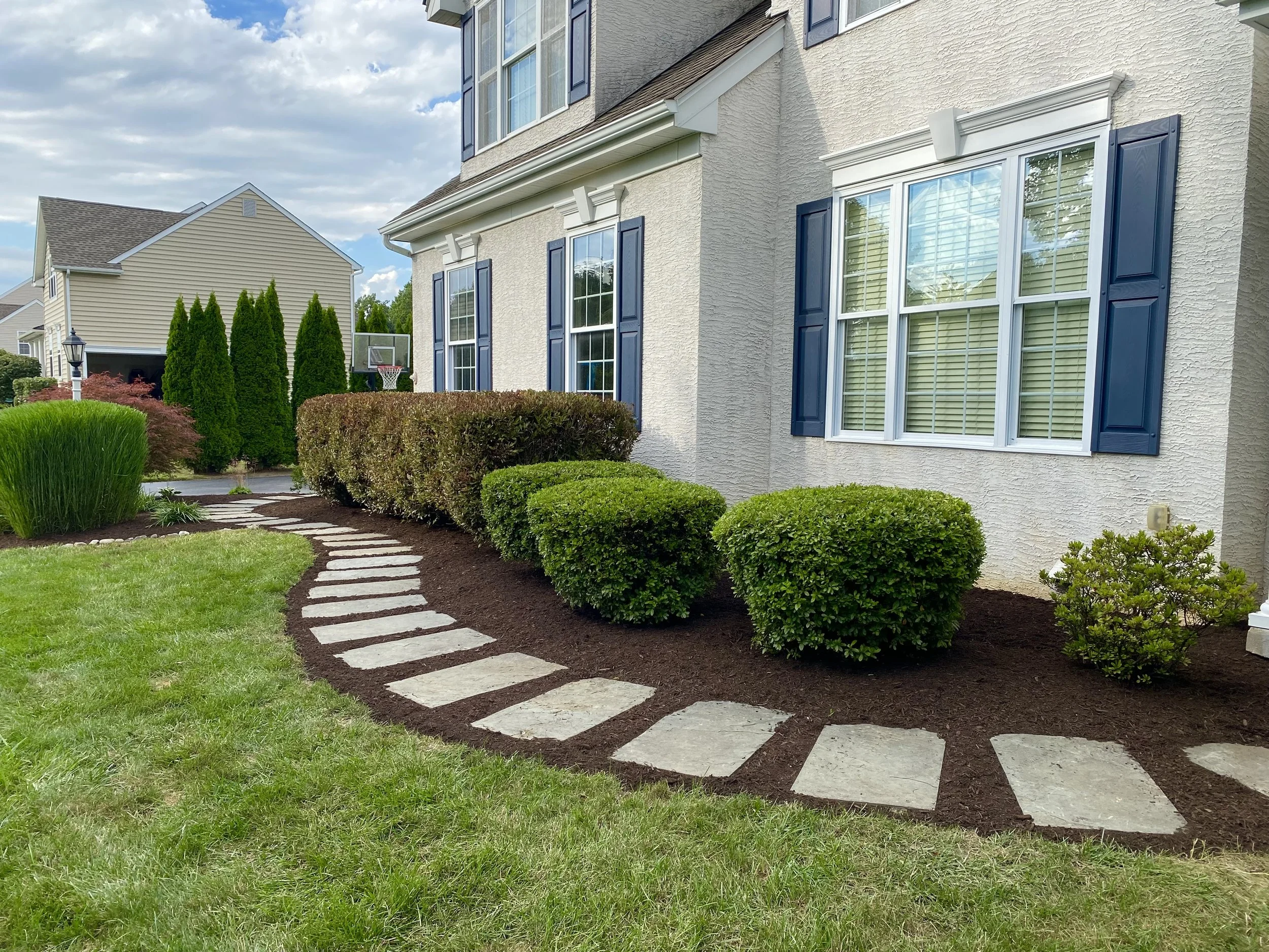 A well-maintained front yard with a curved stone pathway leading to a house with beige siding and blue shutters. The yard features neatly trimmed bushes, green grass, and a cloudy sky.