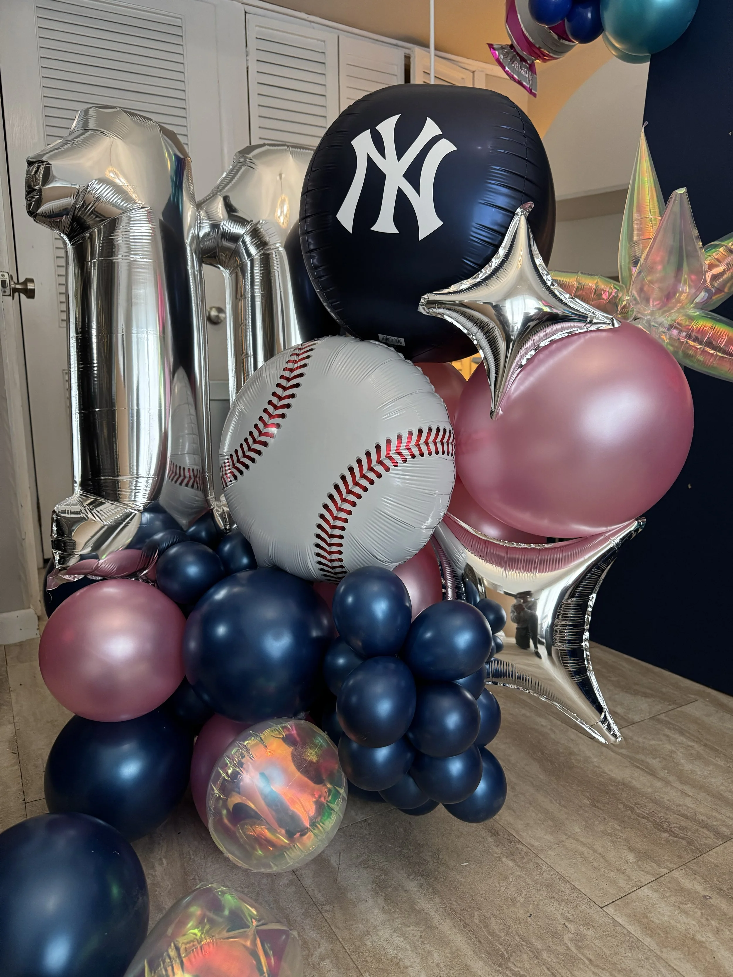 Balloon arrangement with a large black New York Yankees baseball, a large white baseball with red stitching, pink and dark blue balloons, silver star-shaped balloons, and a silver number one balloon, for a baseball-themed celebration.