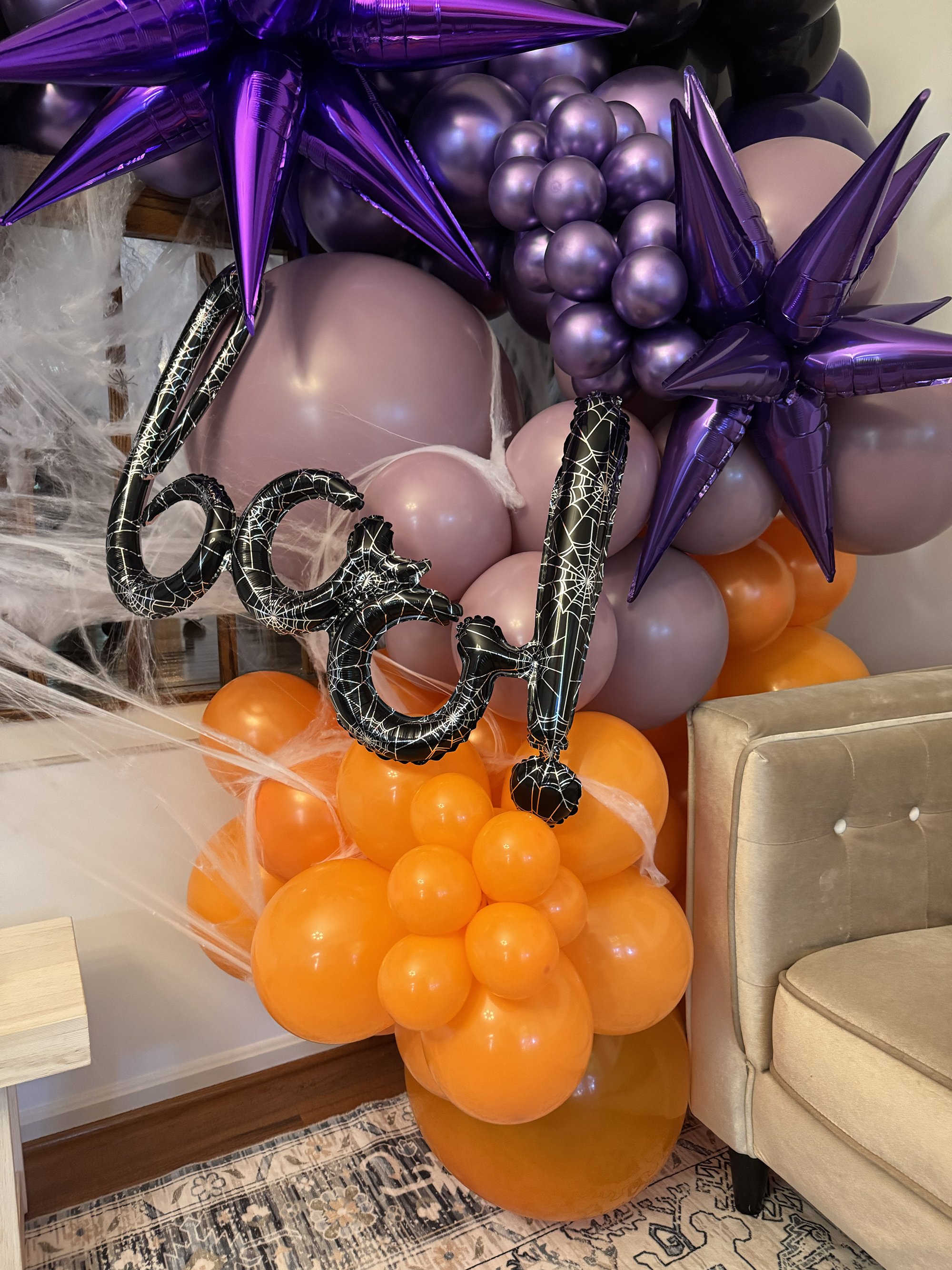 A Halloween-themed balloon arrangement featuring purple, orange, and black balloons, with some shaped like stars and others like clusters of circles. There are black balloons with spider web patterns spelling out "BOO!" and black and purple star-shap