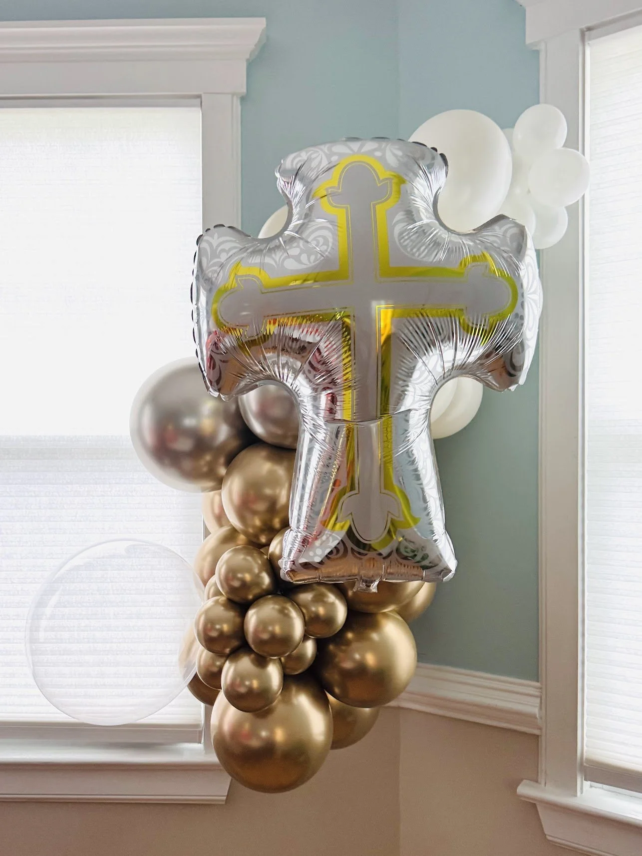 A large metallic balloon shaped like a cross with a yellow outline, surrounded by clusters of gold and white balloons, inside a room with white window blinds and light blue walls.