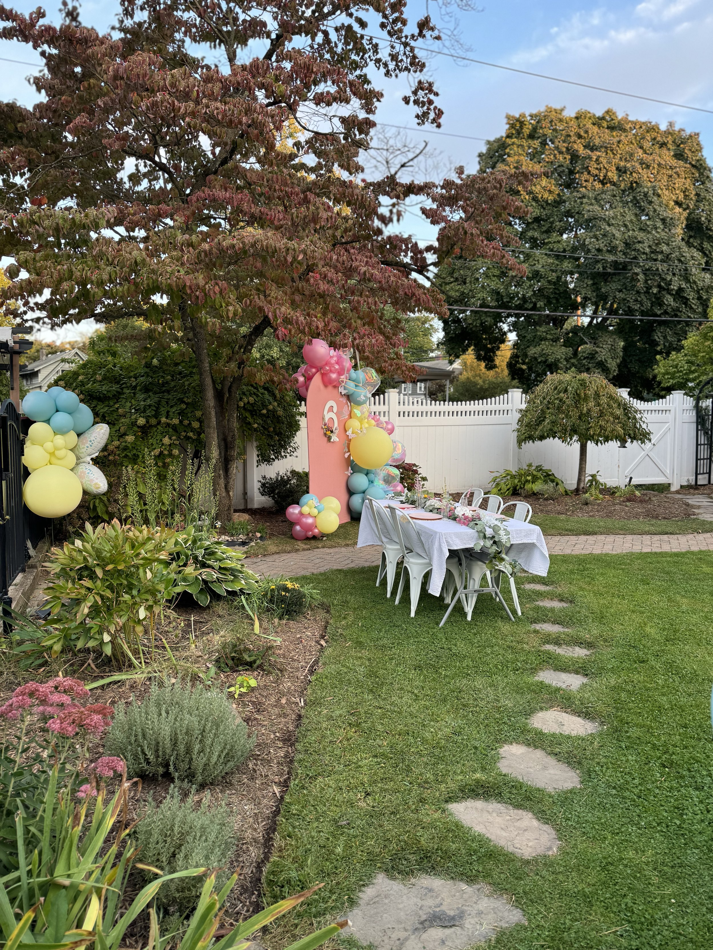 Backyard decorated for a celebration with balloons, a pink arch, a table with a white tablecloth and chairs, a stepping stone pathway, trees, and a white fence.