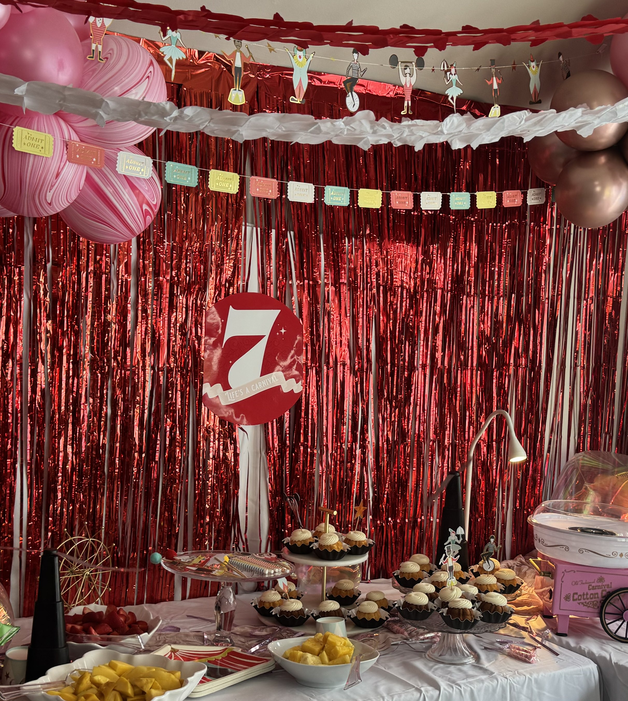 Decorative cupcake table with a red and white theme, featuring a large red circle with the number 7 and the phrase 'Life's a Carnival,' cupcakes on display, and colorful banners and balloons in a party setting.