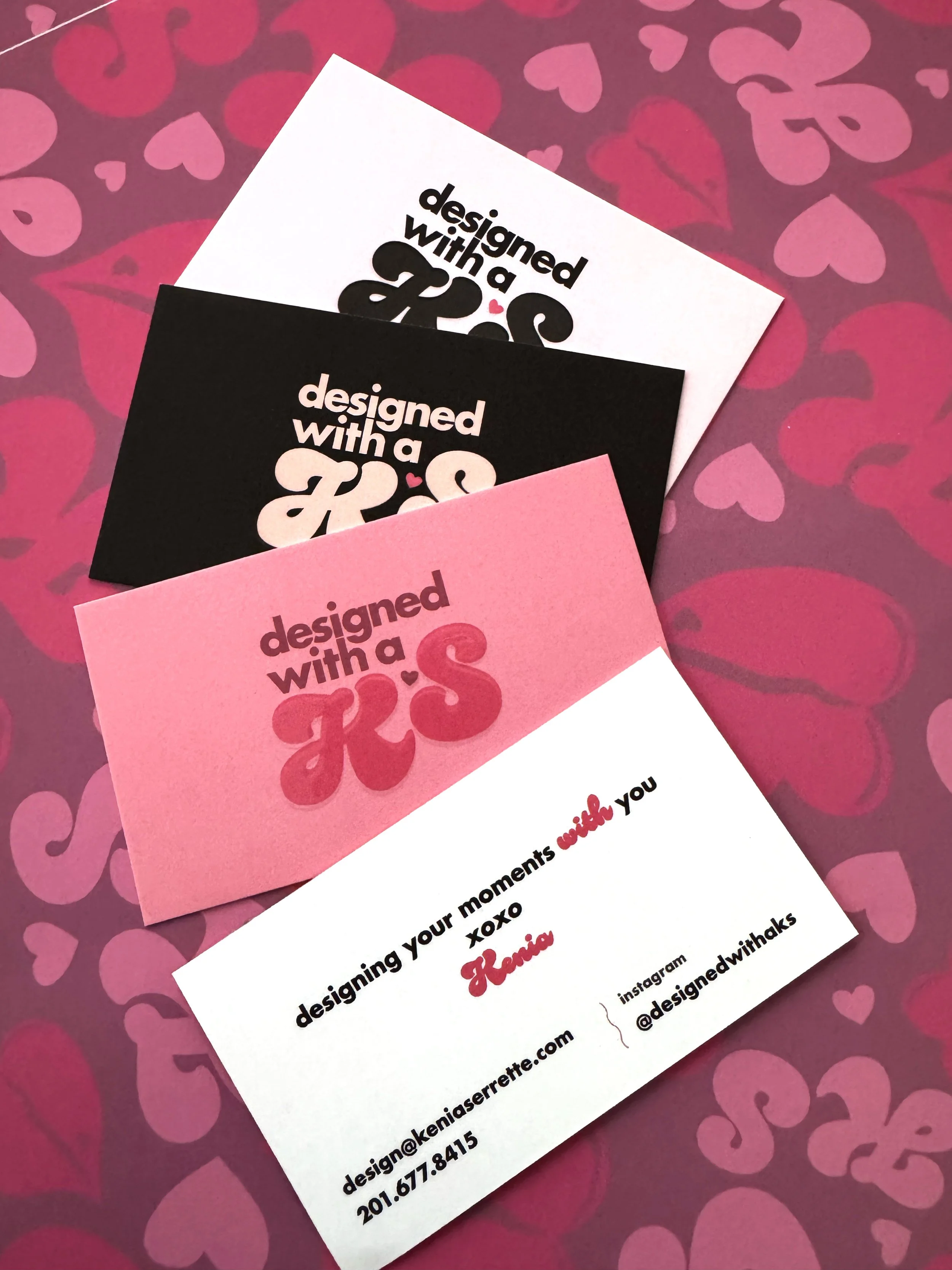 Business cards with the phrase 'designed with a KS' in various colors, placed on a pink and red heart-patterned background.