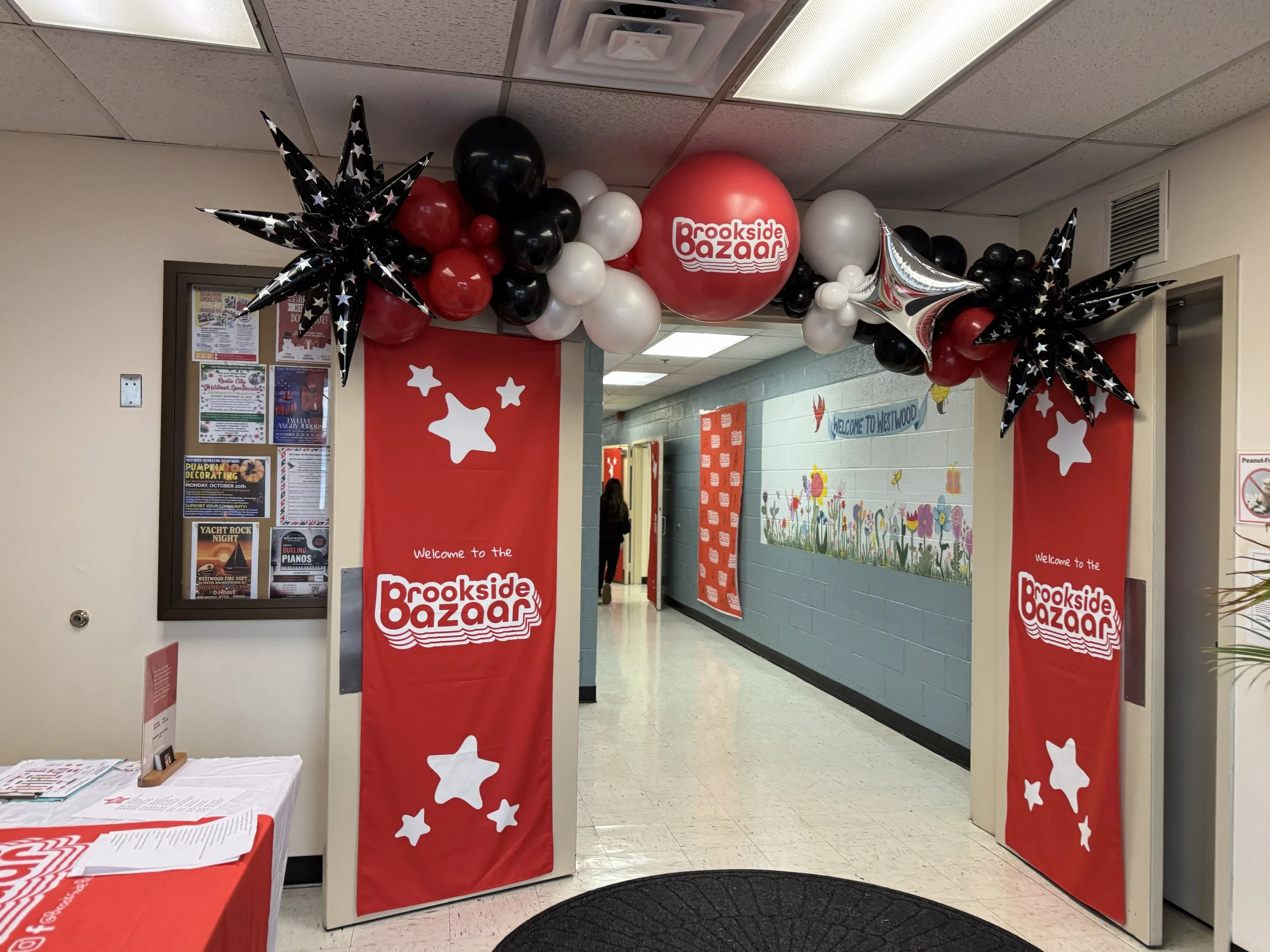 Decorated entrance to Brookside Bazaar with red banners, black, white, and red balloons, and black star-shaped balloons with white stars. A table with flyers is on the left. Inside, the wall has colorful flowers and a sign that says "Welcome to Westw