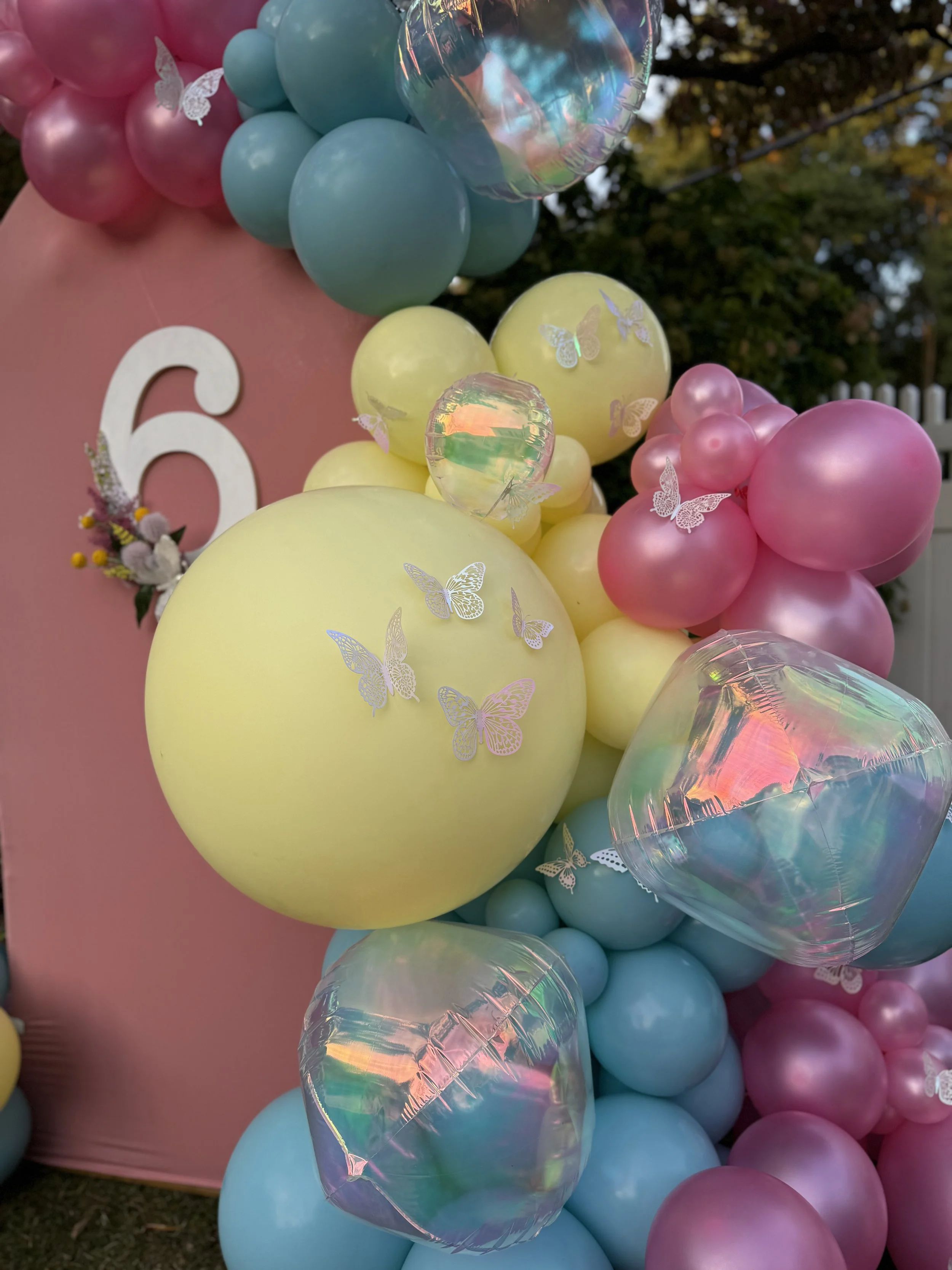 Colorful balloon decoration with pink, yellow, blue, and iridescent balloons arranged behind a pink backdrop with the number 6. Butterfly stickers are on some balloons, and there is greenery and a white fence in the background.
