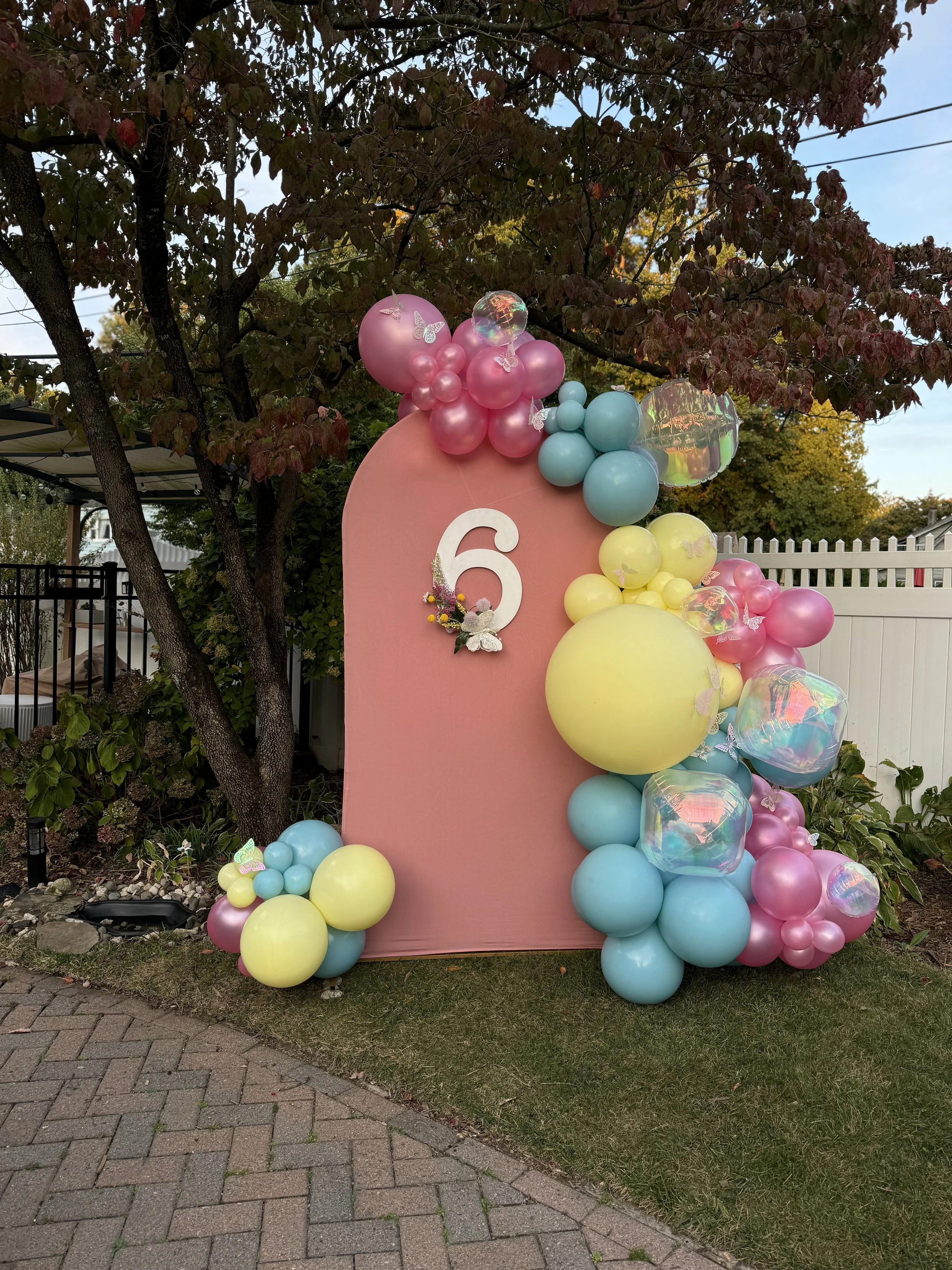 Decorative outdoor birthday setup with pink, yellow, blue, and iridescent balloons arranged around a pink backdrop with the number 6 and small flower and butterfly decorations.