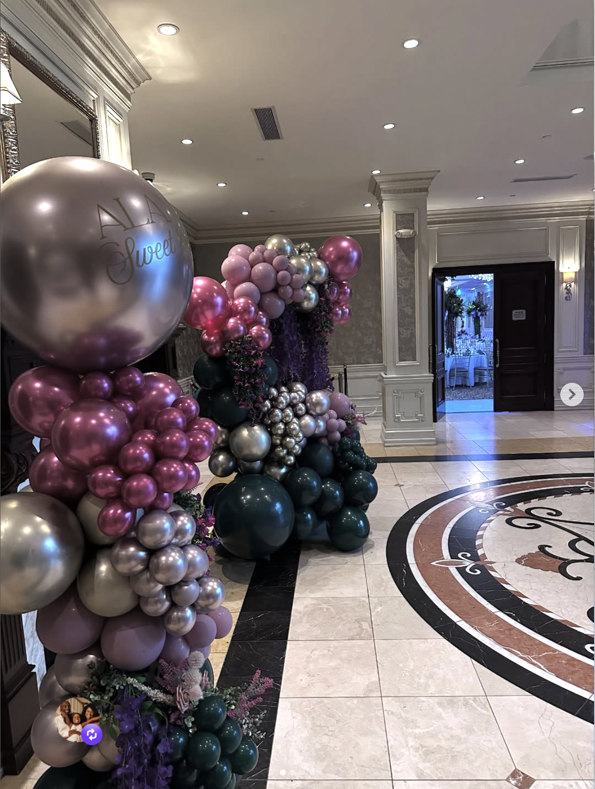 Decorative balloon arch with pink, silver, and dark green balloons at an indoor event venue, with a floral arrangement at the base and an ornate floor design nearby.