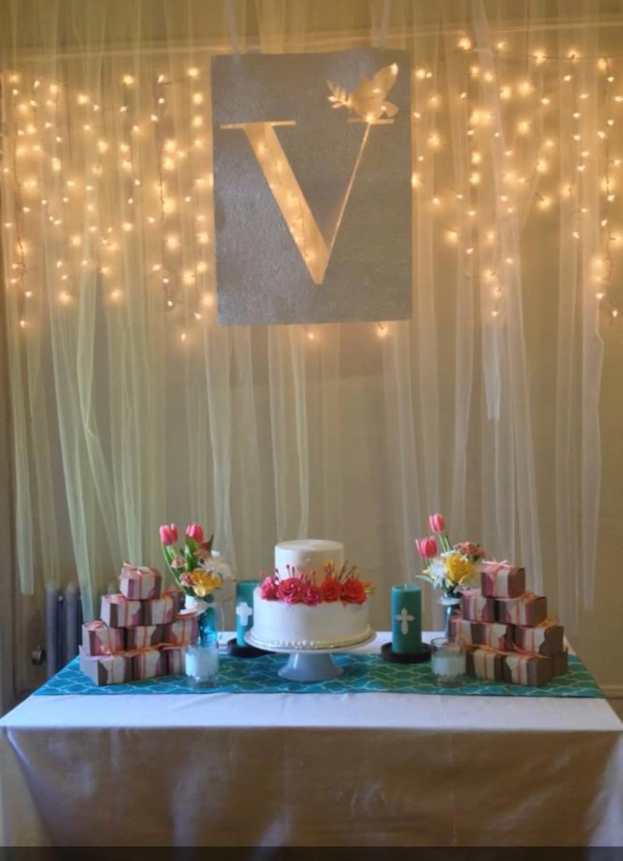 Decorated table with a white cake adorned with pink and yellow flowers, surrounded by wrapped gifts and floral arrangements, set against a backdrop with hanging string lights and a large silver letter 'V' with a butterfly and leaves design.