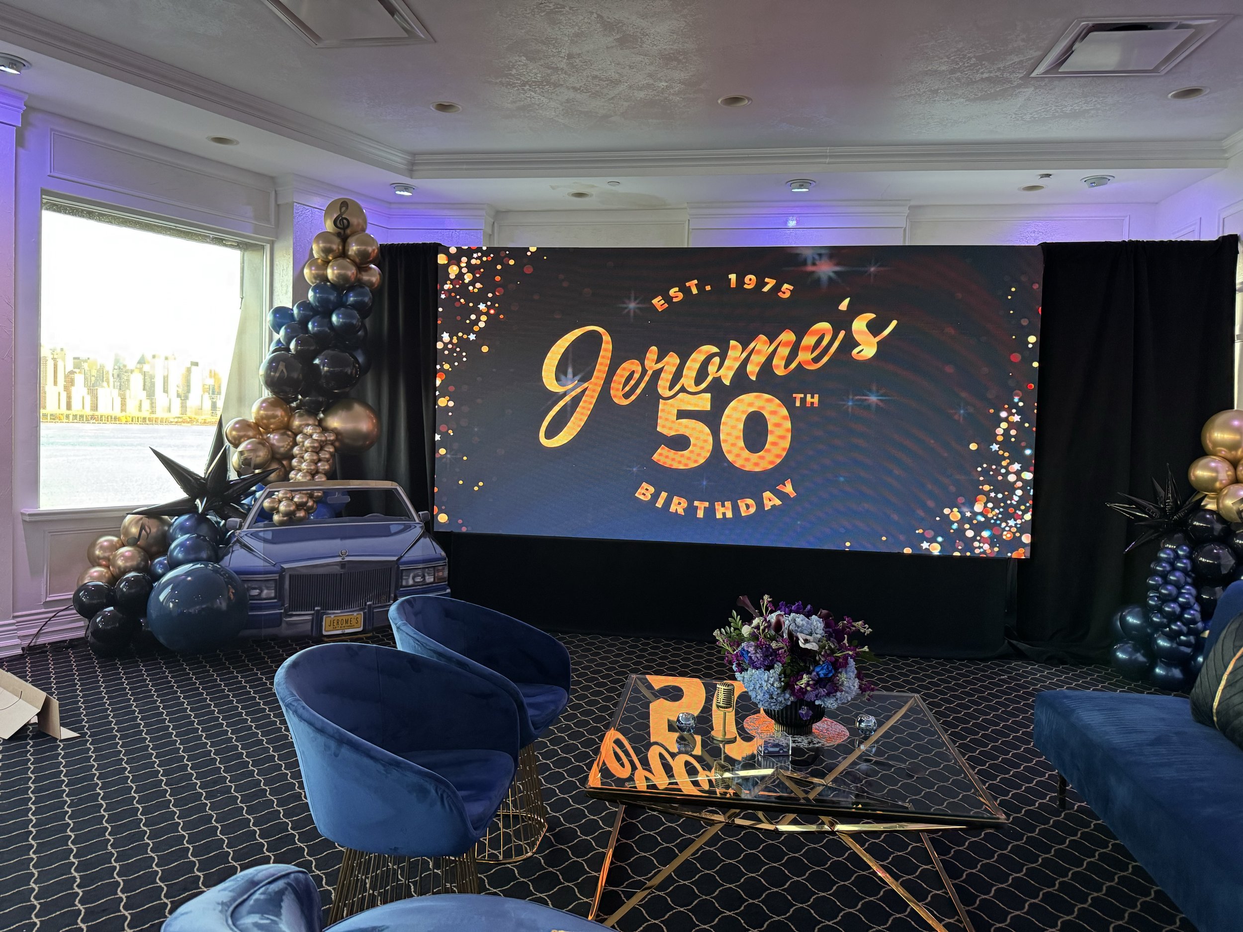 Indoor celebration decor for Jerome's 50th birthday, with a large digital screen displaying the event name and anniversary, balloons in black, gold, and purple, a prop car with a license plate and balloons, and blue velvet chairs and a glass table with floral centerpiece.