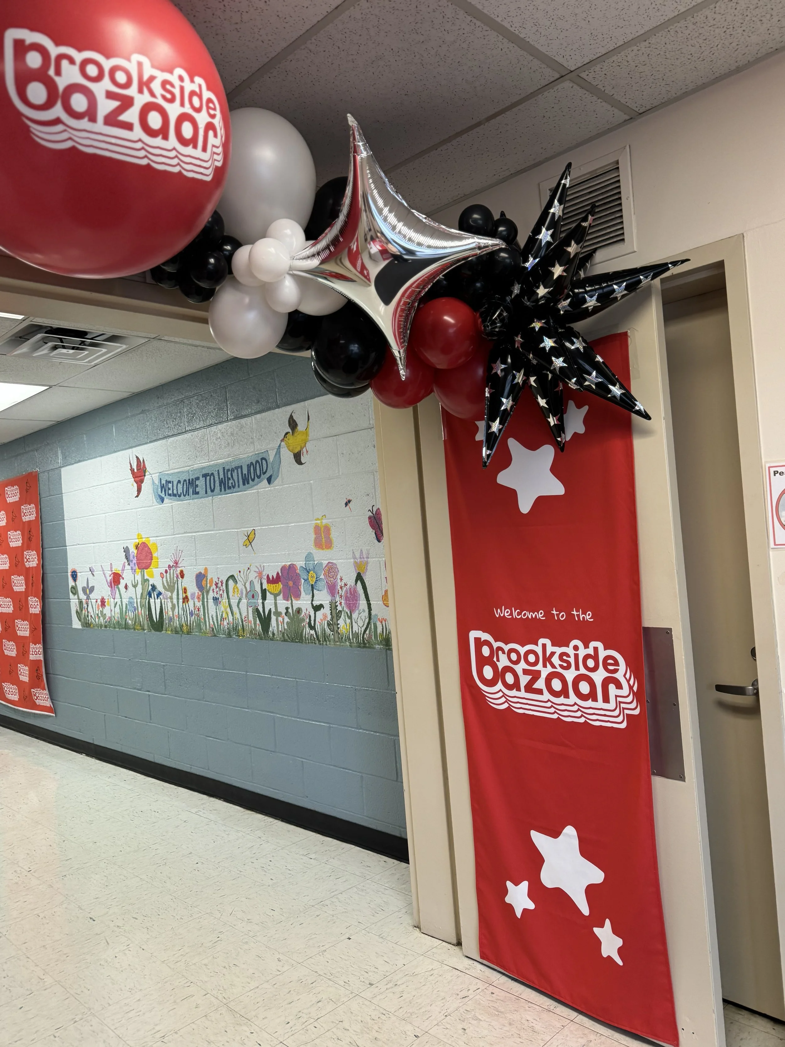 Decorations for the Brookside Bazaar, including a large red balloon with the event name, a cluster of smaller black, white, and red balloons, and black star-shaped balloons with white stars, hanging in an indoor hallway with a mural of colorful flowe