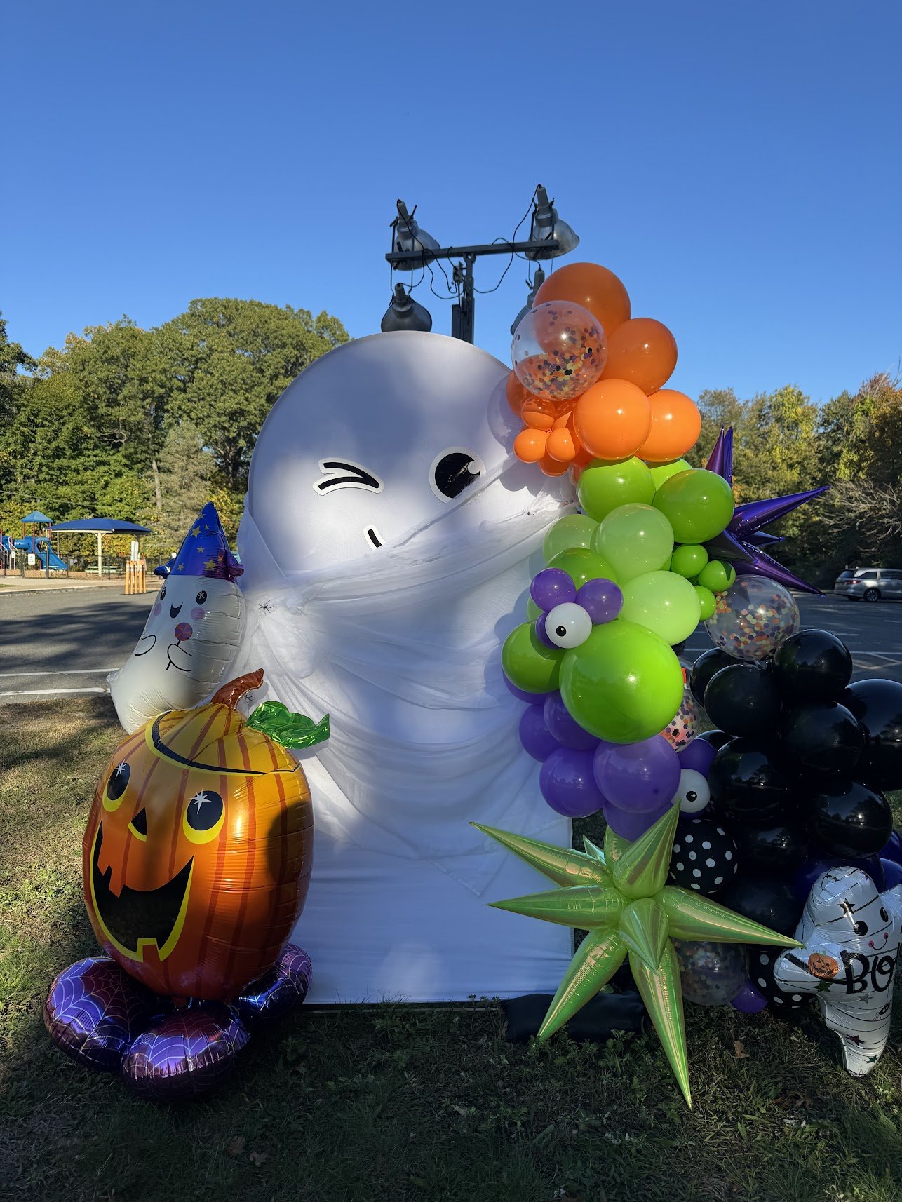 Halloween-themed balloon display with a ghost face, pumpkin, witches hat, colorful balloons, and spooky decorations outdoors on a sunny day.