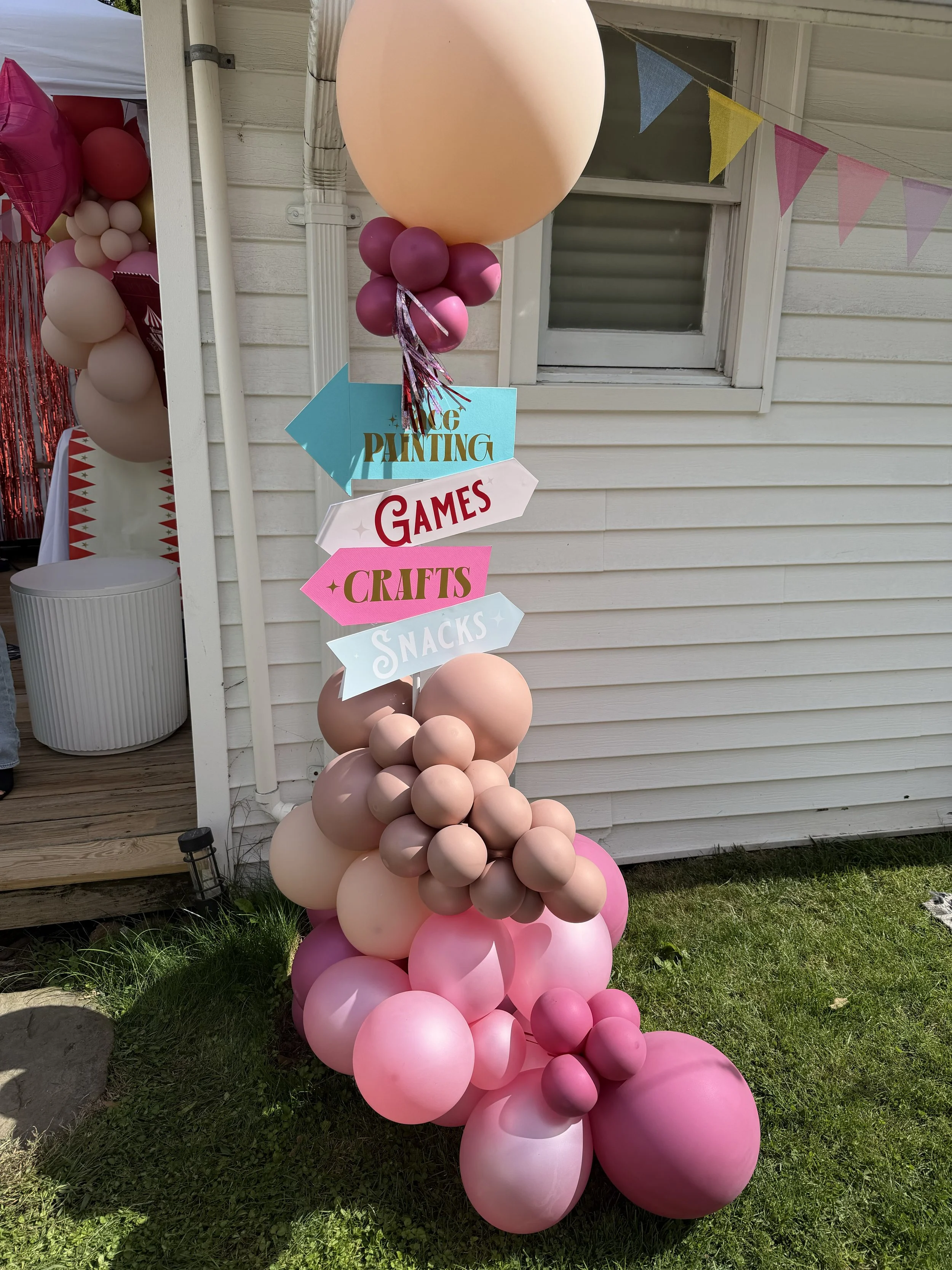 Decorative balloon column with pink, peach, and purple balloons, with directional signs for face painting, games, crafts, and snacks, outside a house with white siding.