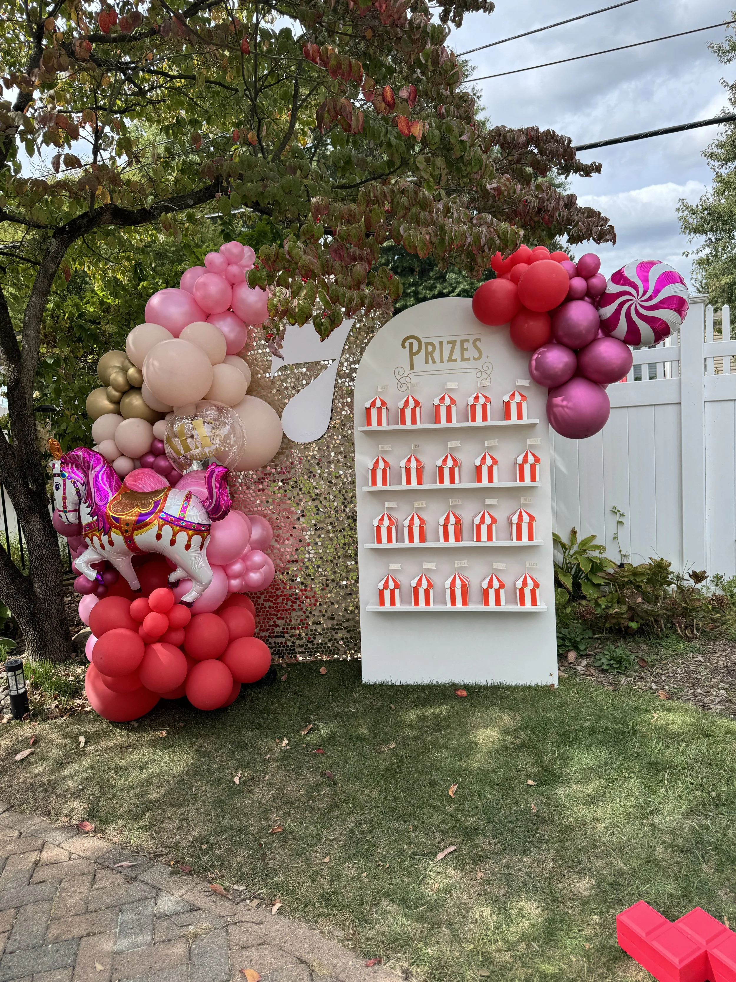 Decorative prize display with striped tent-shaped containers, pink and red balloons, a carousel horse balloon, and a golden sequin backdrop, set outdoors near a tree and white fence.