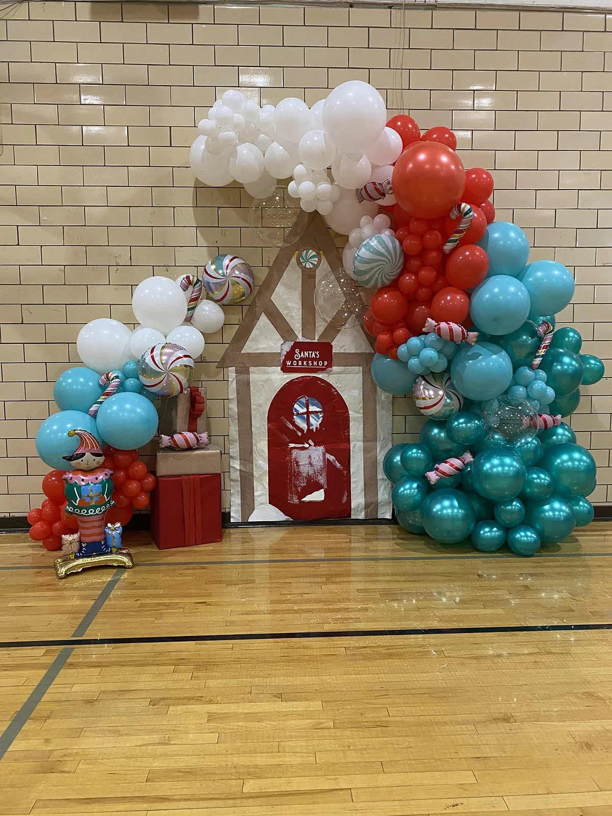 Festive holiday display with a paper house, colorful balloons in red, white, and teal, striped candy canes, wrapped presents, and an elf figure, all arranged against a brick wall.