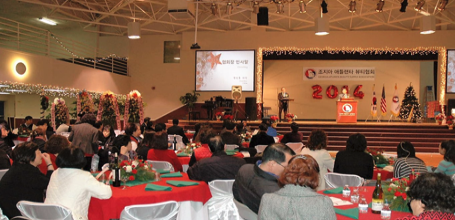 A large indoor event with a stage, decorated with Christmas trees and lights, where people are seated at tables attending a presentation or celebration.
